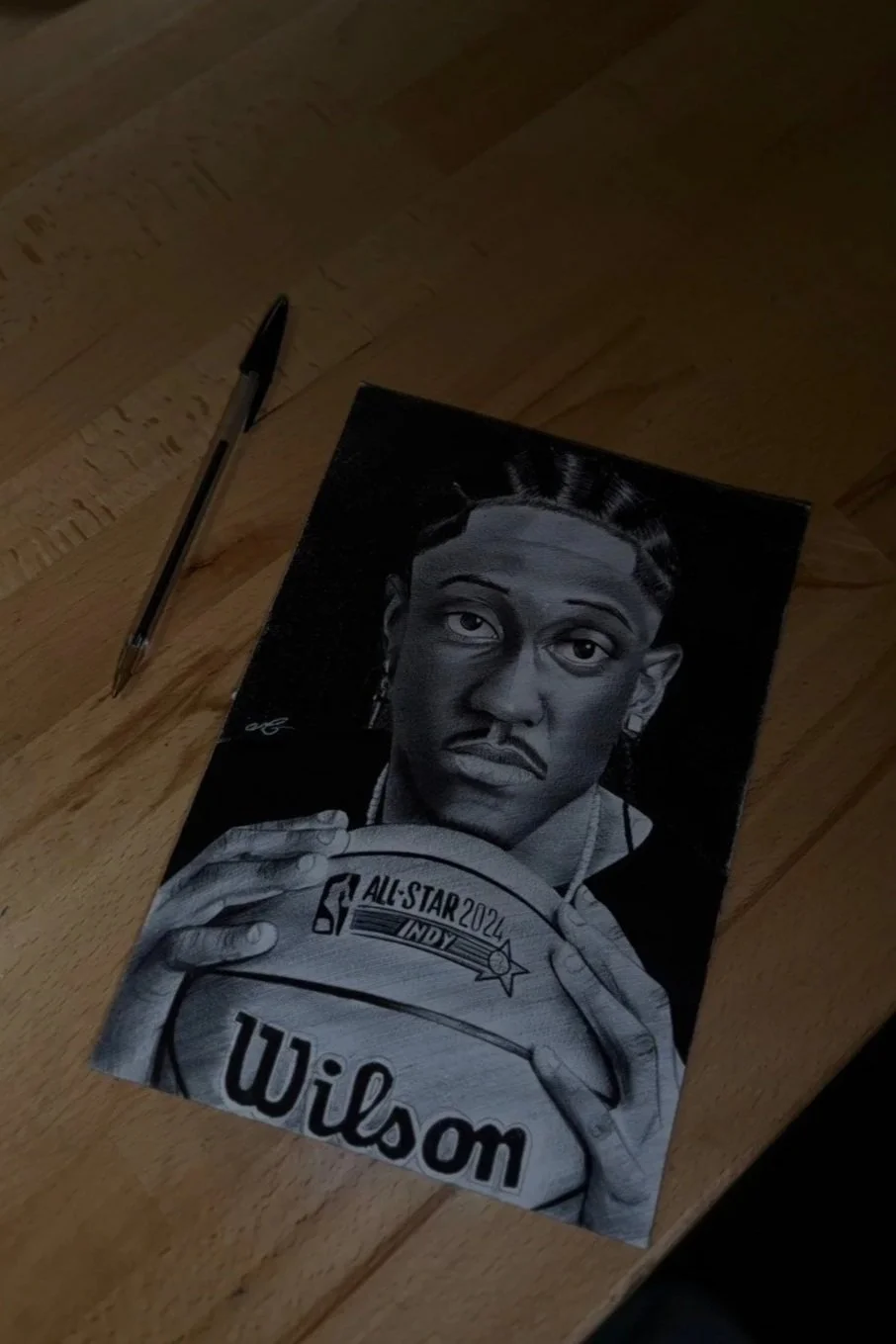 Black and white drawing of a woman holding a basketball with an 'All-Star 2024 INDY' logo, and the name 'Wilson' at the bottom. A pen is placed next to the drawing on a wooden surface.