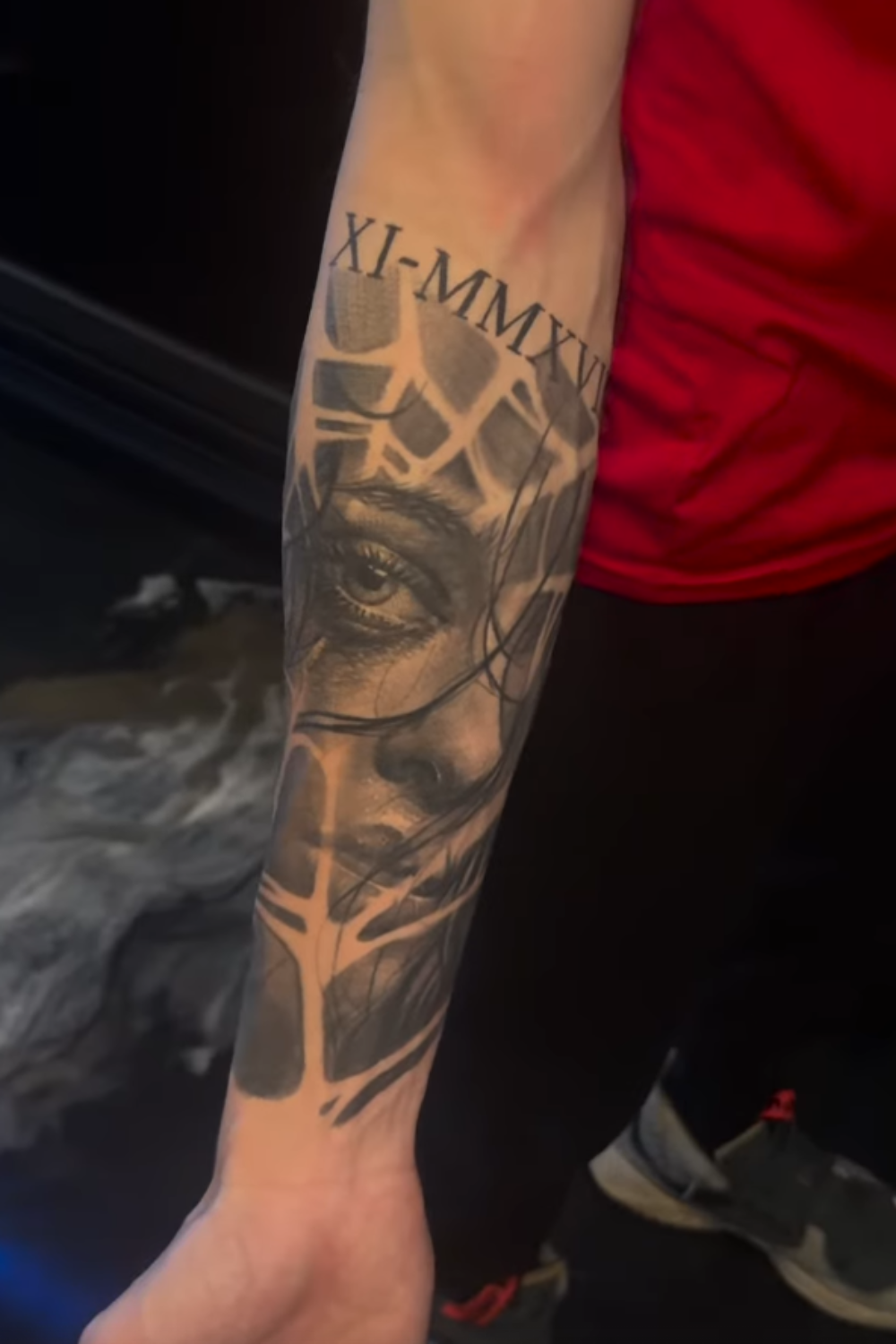 Tattoo of a realistic eye and part of a human face on a person's forearm, with Roman numerals above.