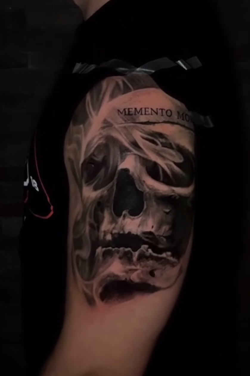 A tattoo of a skull with detailed shading, featuring a banner with the Latin phrase "MEMENTO MORI" across the forehead.