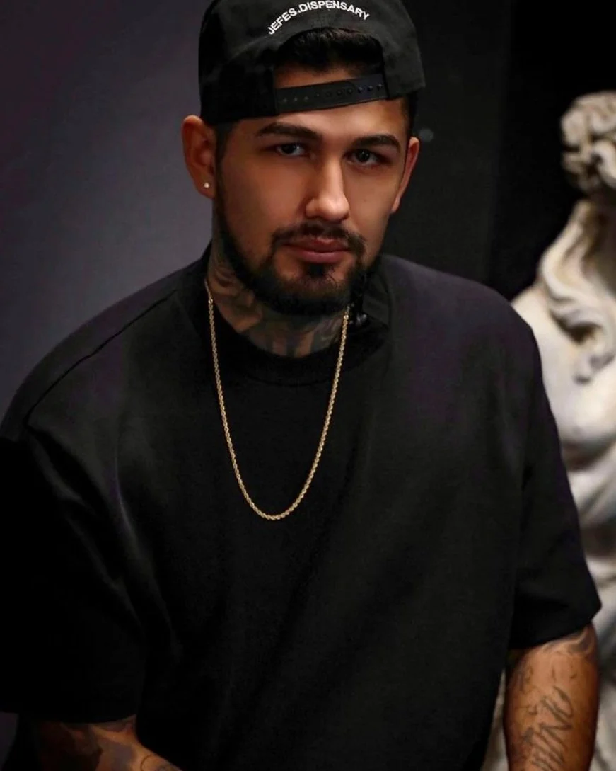 A young man with a beard and tattoos on his neck and arms, wearing a black cap backwards and a gold chain, looking at the camera.