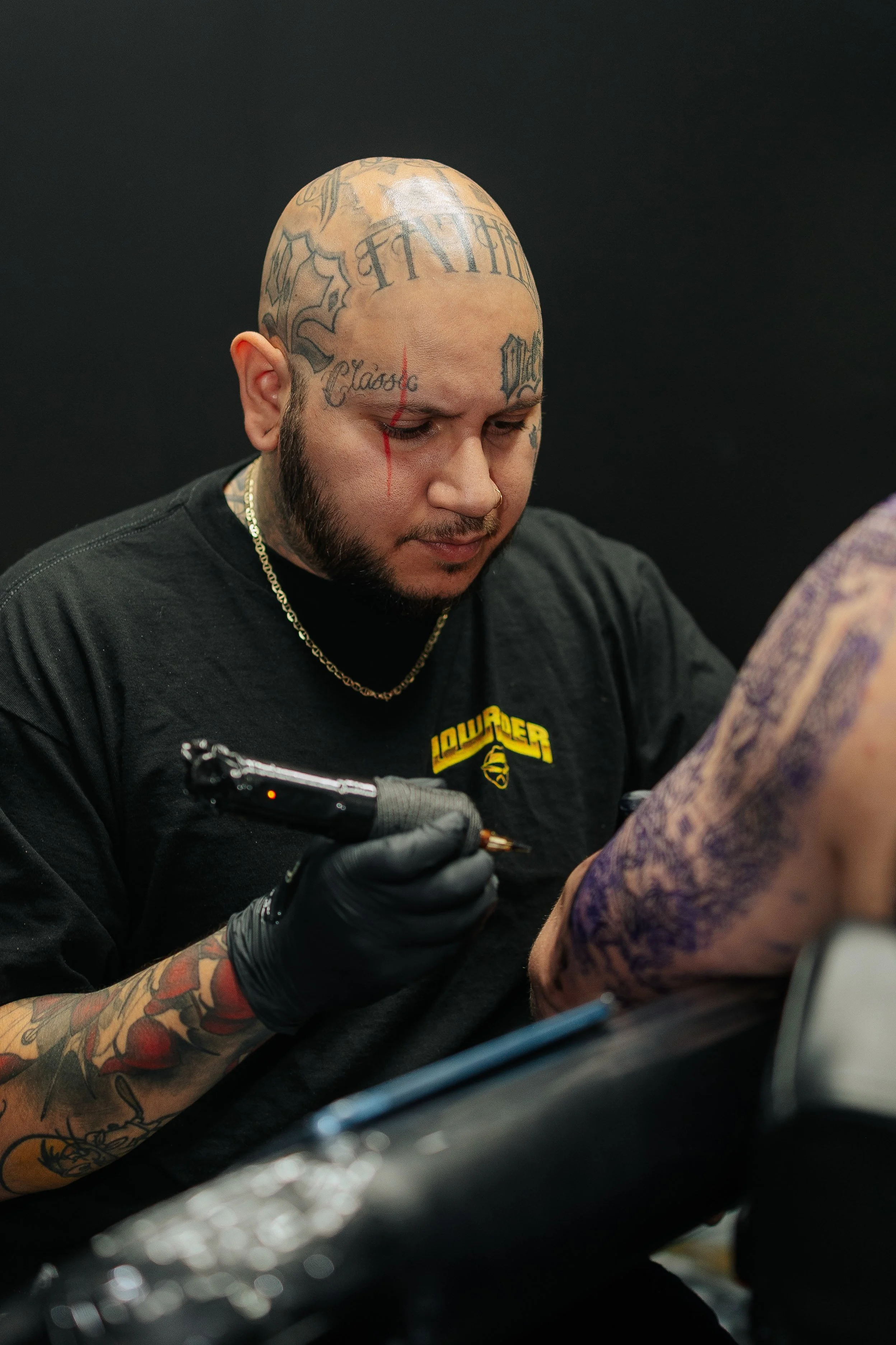 A tattoo artist with tattoos on his head and arm, wearing a black t-shirt with the word 'LOWBLOW' and a logo, is using a tattoo machine on a person's forearm, against a black background.