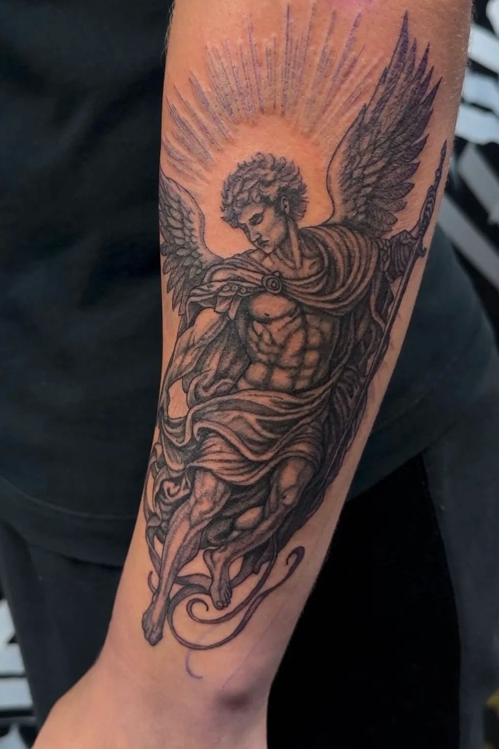 Tattoo of an angel with large wings, a muscular torso, and a halo, holding a sword, with rays behind the head.