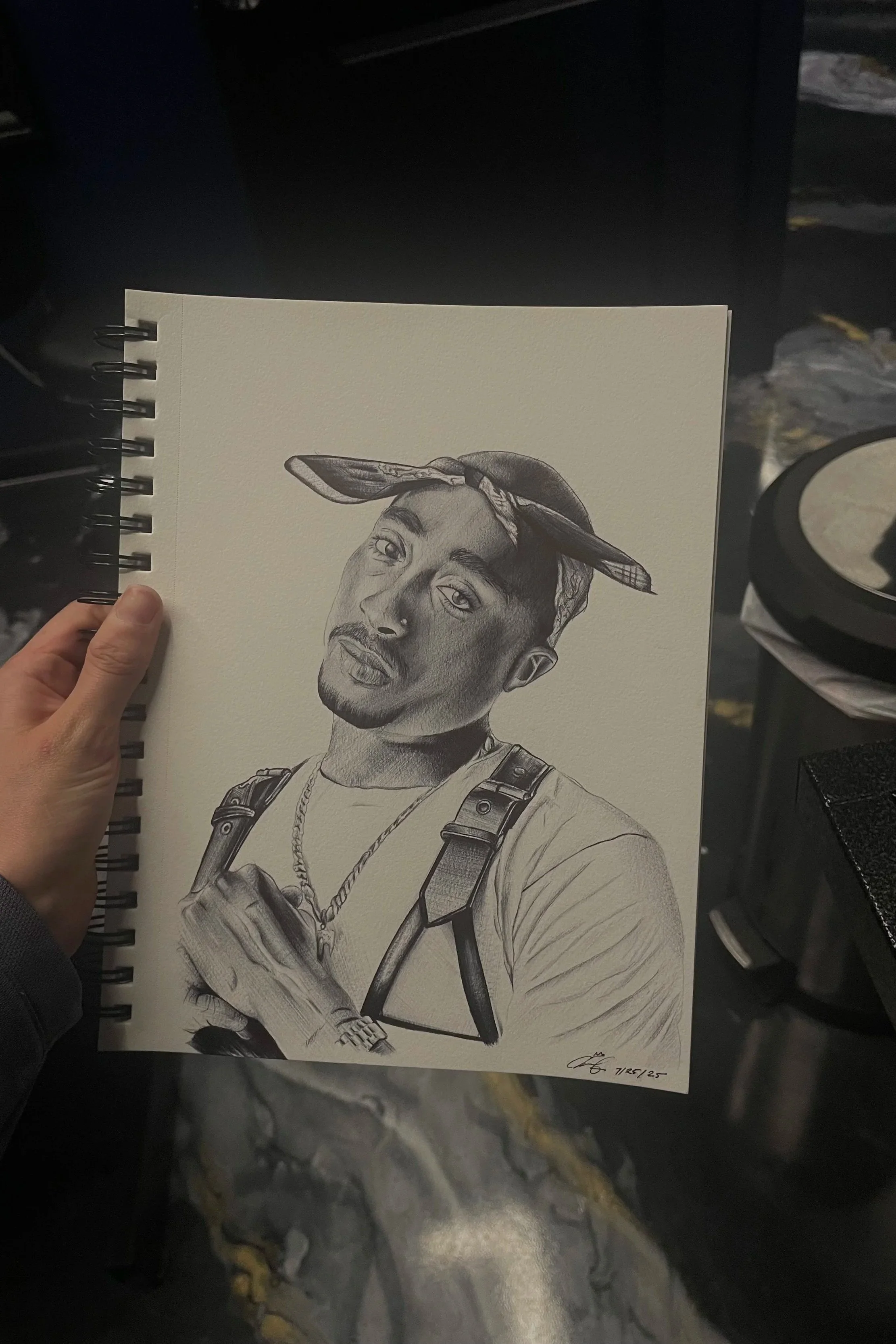 A hand holding a spiral-bound sketchbook with a detailed black-and-white portrait of a young man with a bandana on his head. The portrait depicts the man with a serious expression, wearing a necklace and a harness. The sketchbook is shown against a dark background with a marble or stone surface underneath.