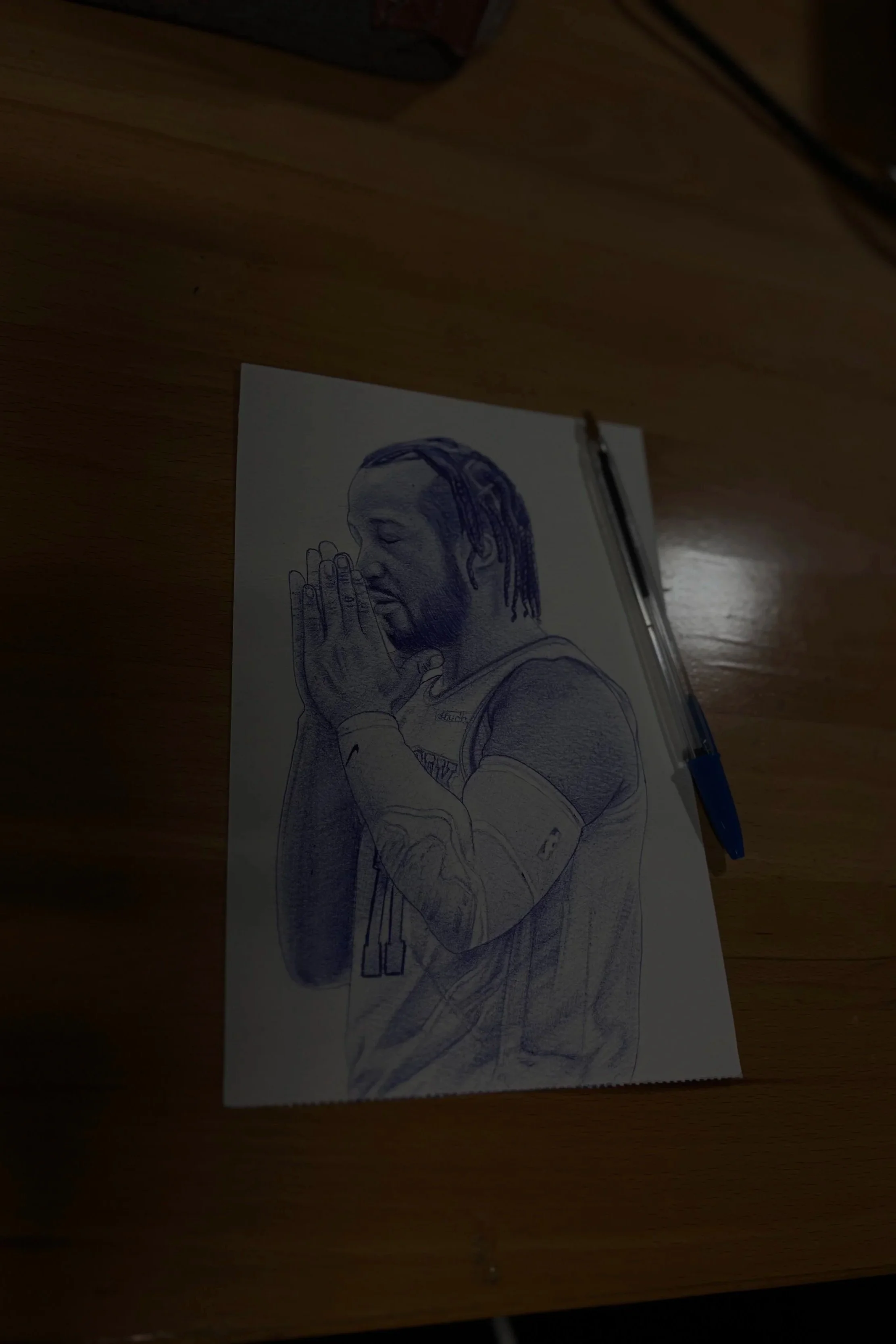 A detailed pencil sketch of a person with dreadlocks and hands pressed together in a praying or meditative pose, wearing a long-sleeved shirt, on a piece of paper on a wooden surface with a pen placed beside it.