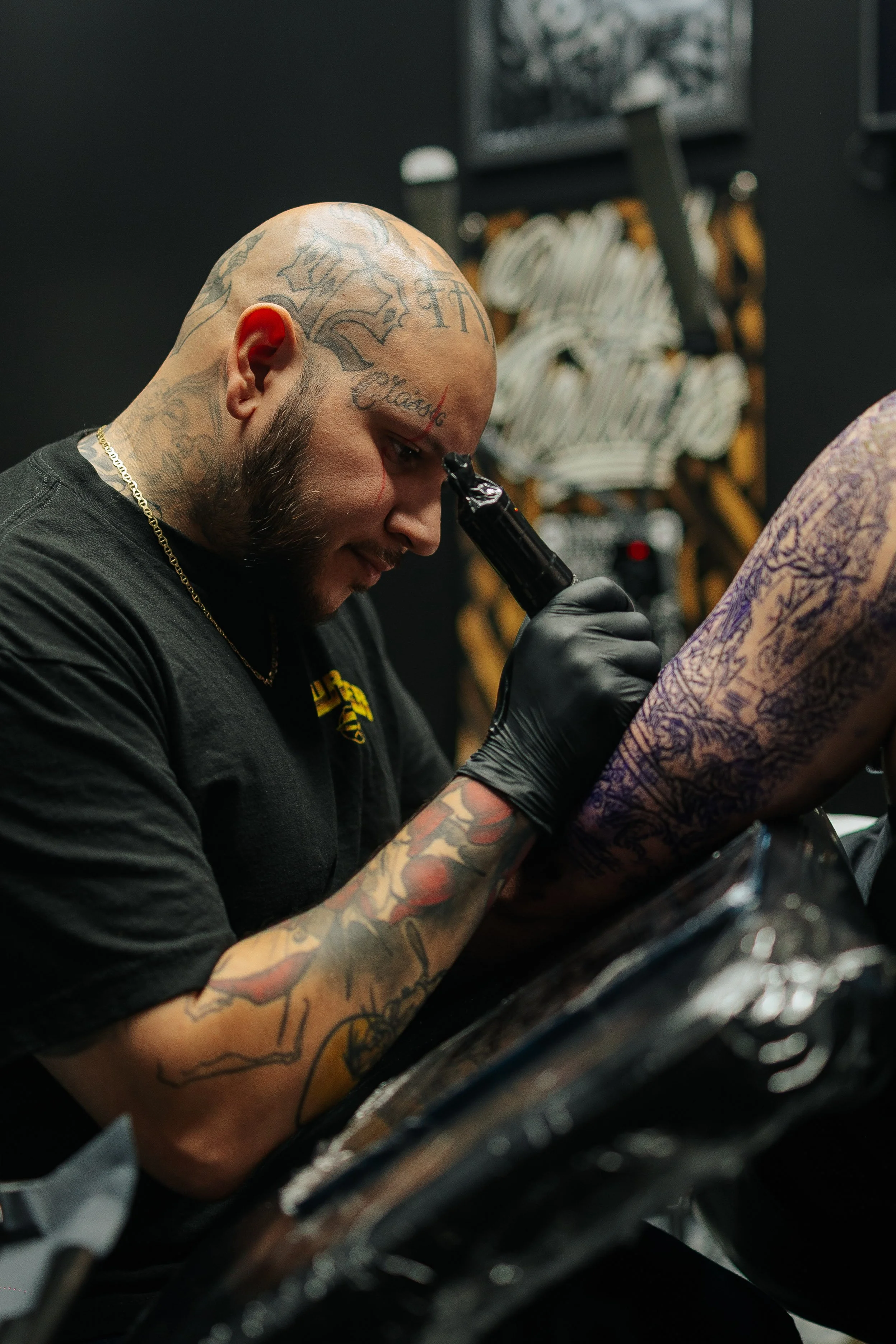 A tattoo artist with tattoos on his head, arms, and face, wearing black gloves, creating a large tattoo on a client's arm in a tattoo studio.
