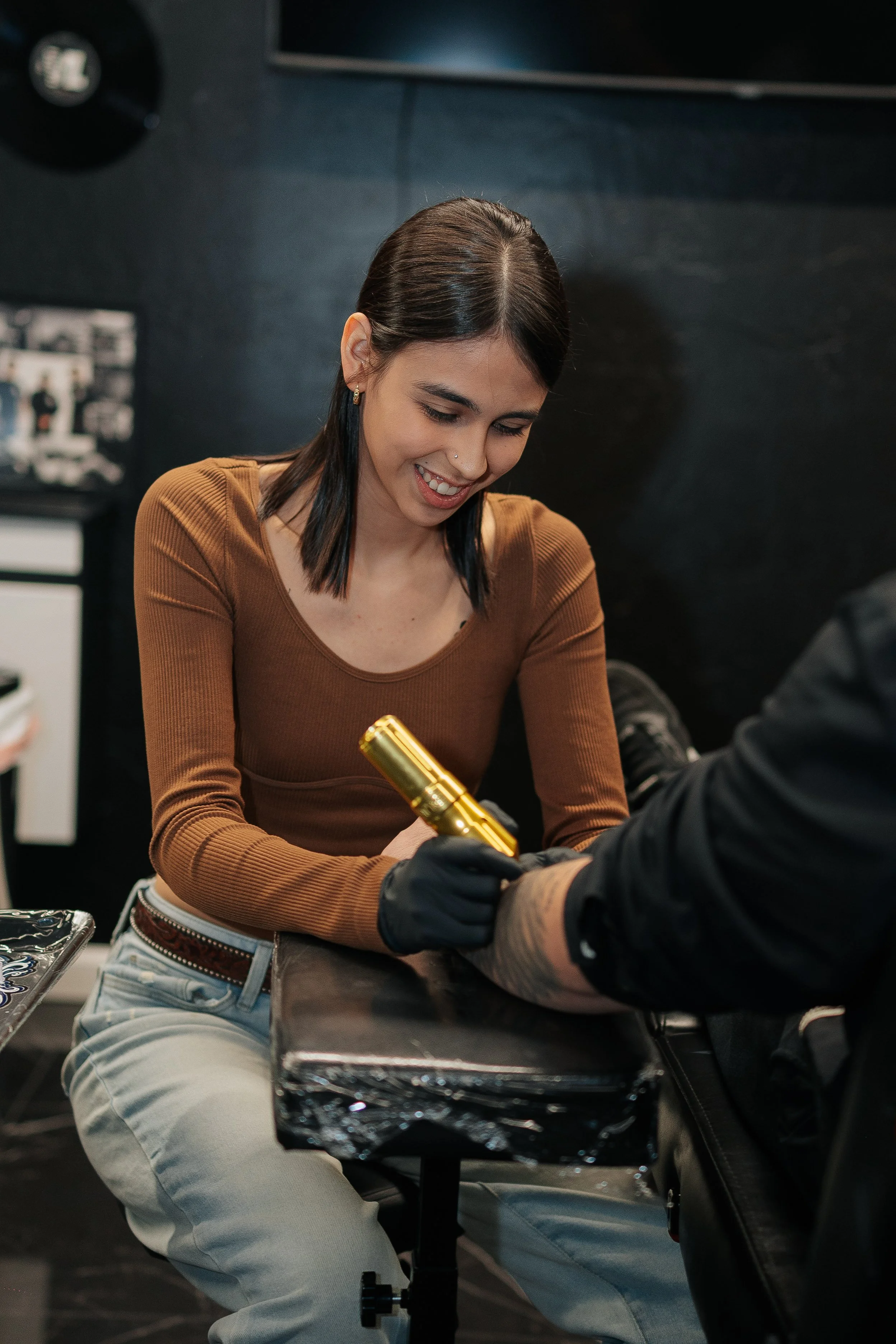 A woman with dark hair and earrings appears to be getting a tattoo on a person's arm in a tattoo studio. She is smiling and holding a tattoo machine with a golden barrel.