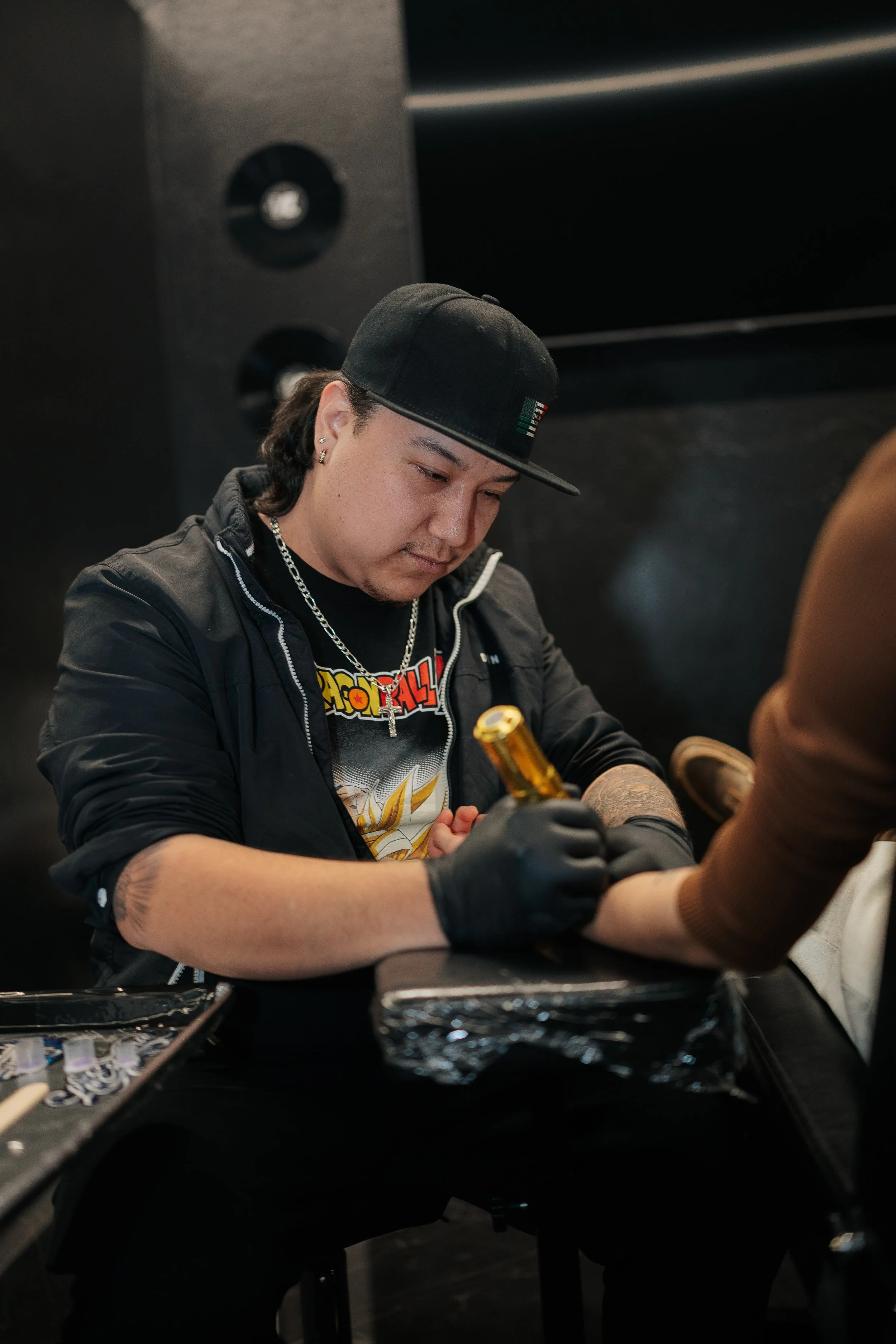 A man with long dark hair, wearing a black cap, black jacket, and jewelry, is giving a tattoo to another person, whose arm is visible. The man appears focused on his work in a tattoo studio.