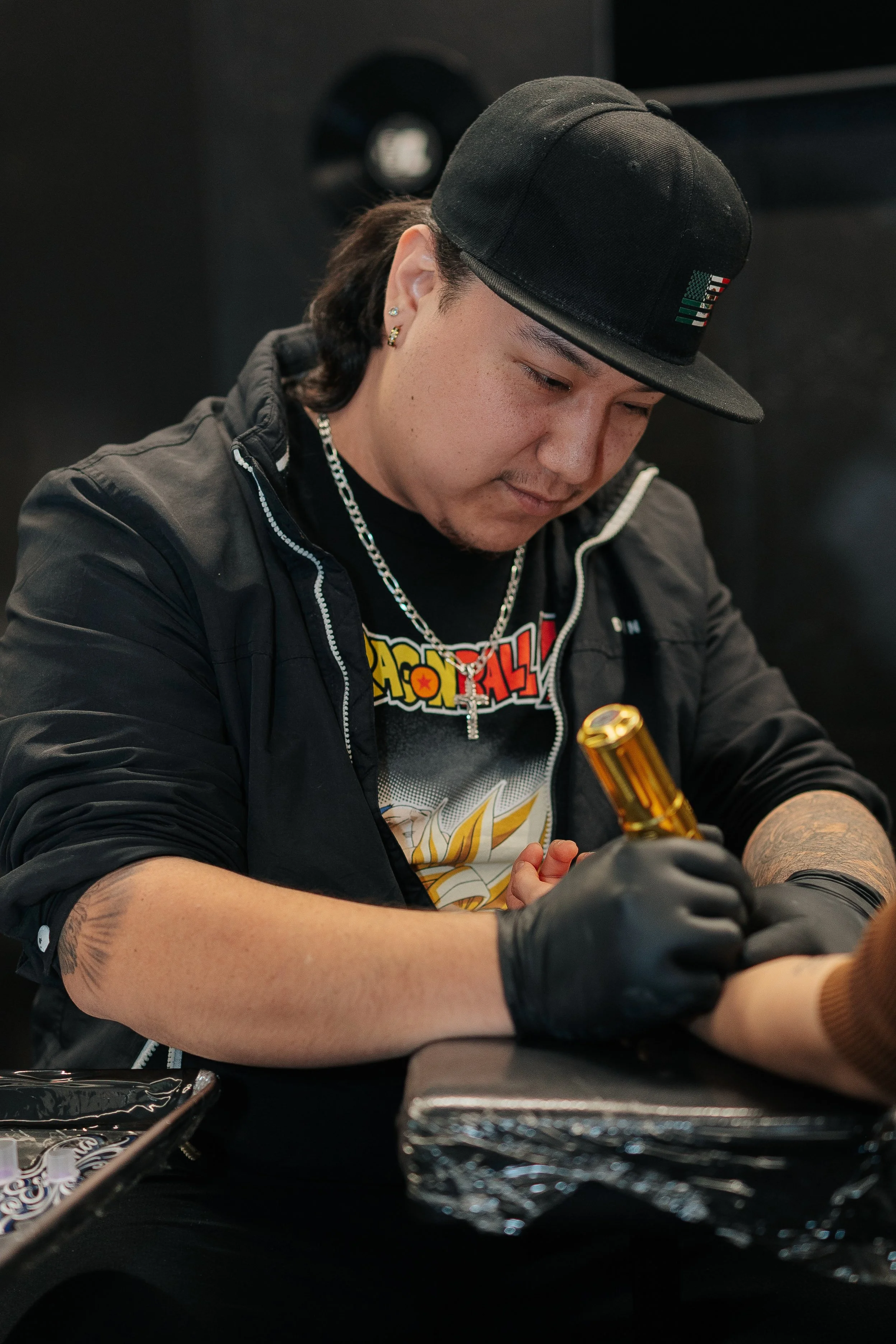 A tattoo artist creates a tattoo on a person's arm. The artist is wearing a black cap, black gloves, a colorful graphic T-shirt, and a chain necklace.