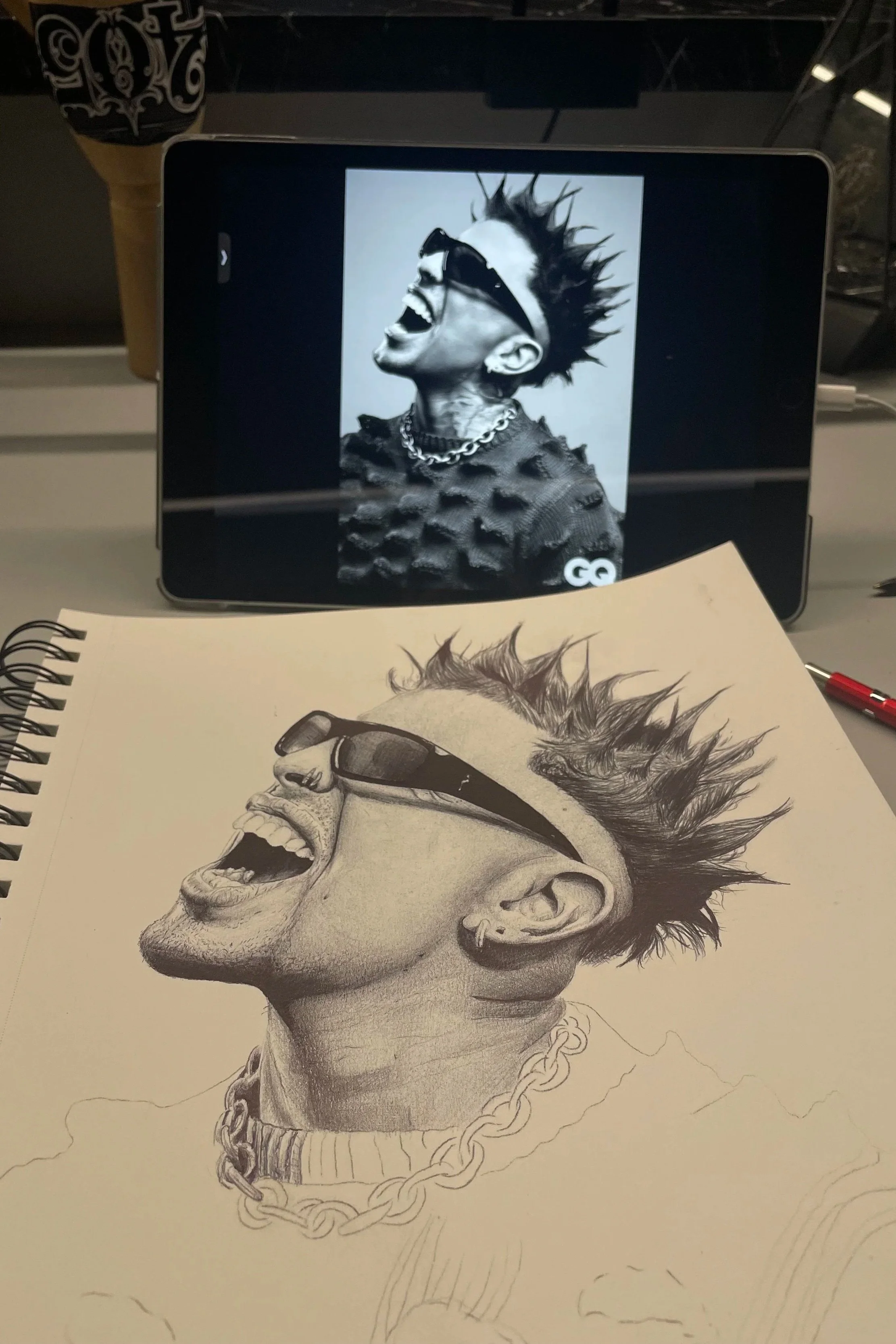 A printed pencil drawing of a man with spiked hair, wearing sunglasses, a chain necklace, and a hoodie, depicted in a lively, expressive pose. A digital device displays a black-and-white photo of the same man in the background.