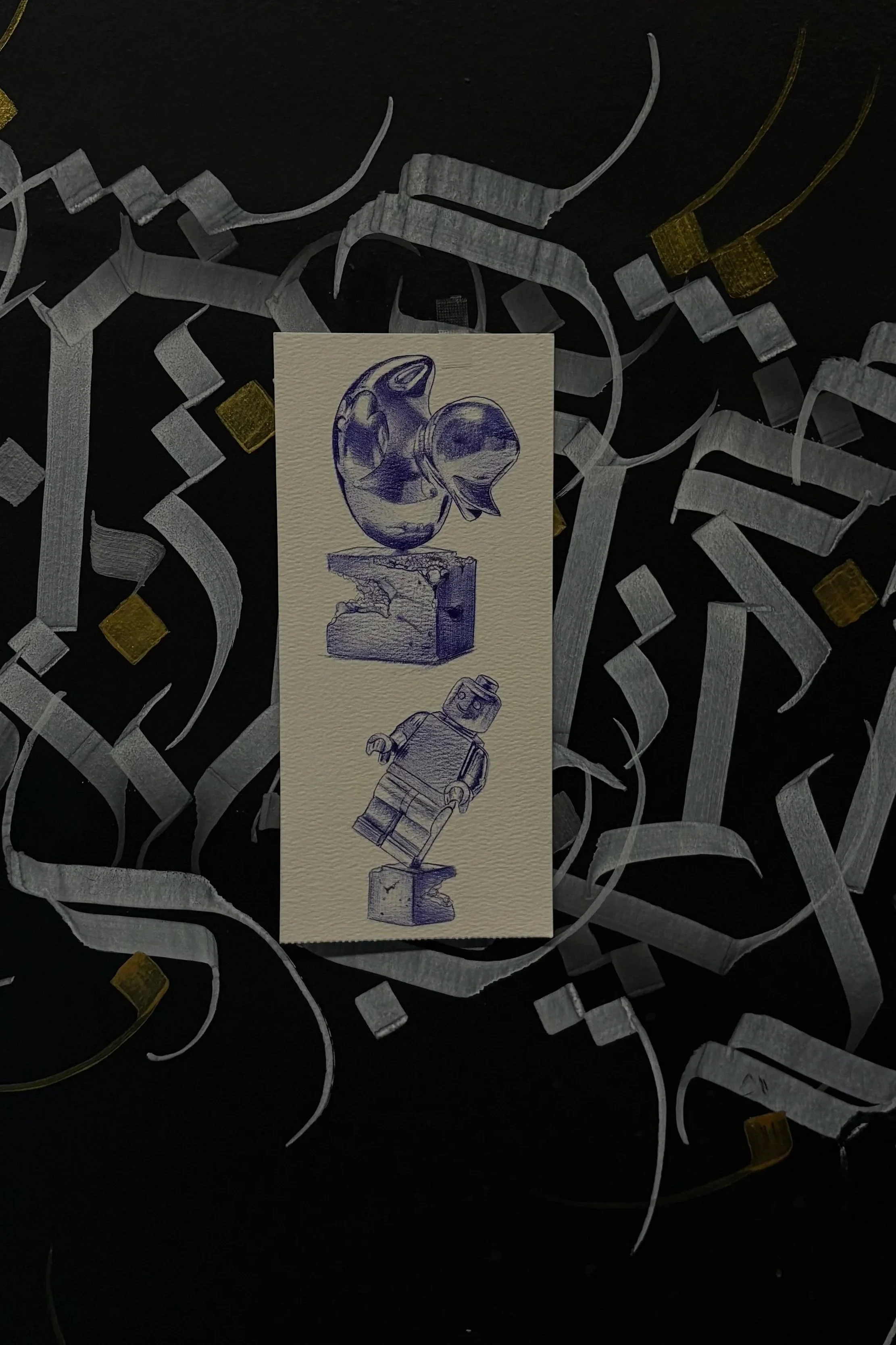 A sketch of a robot and a satellite on a textured white card placed on a dark surface. The background has metallic ribbons and small yellow square confetti.