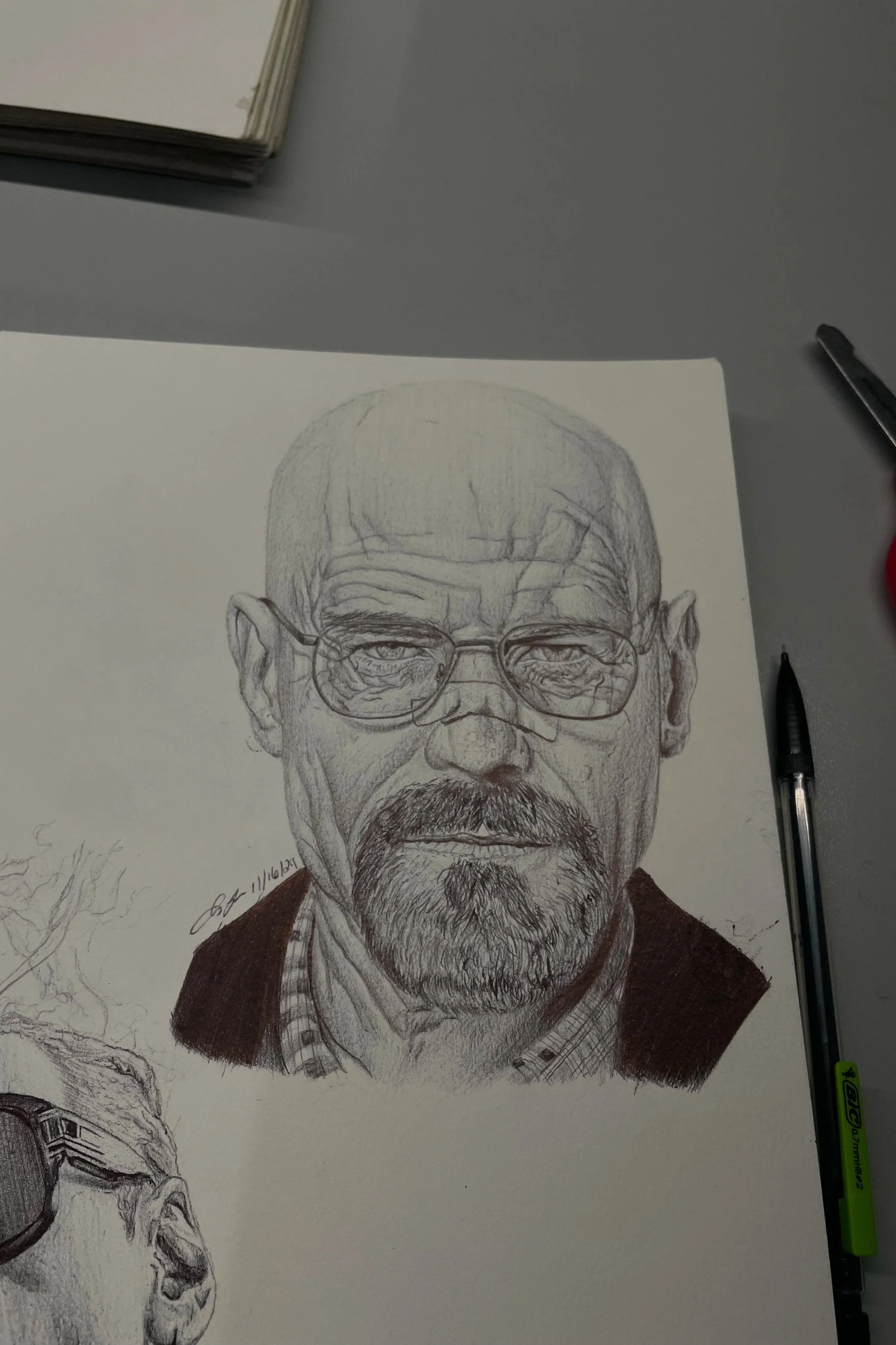 A detailed pencil sketch of an older man with glasses, a beard, and a serious expression, on a piece of paper placed on a gray surface, with a black marker and a green highlighter nearby.