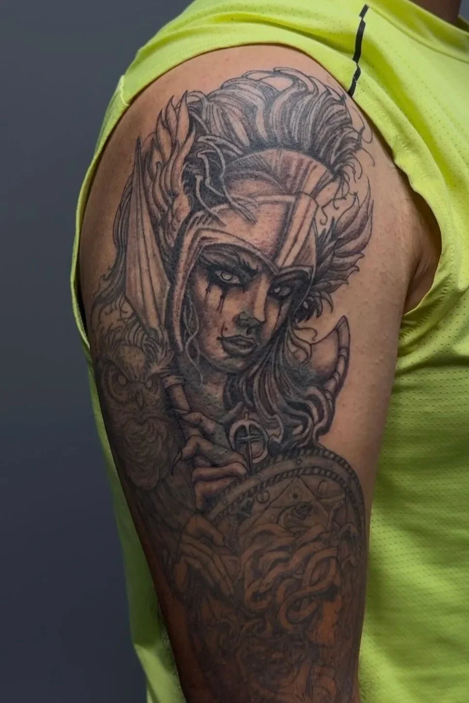 Black and gray tattoo of a fierce woman with wild hair, holding a sword, with intricate details on the arm and circlet, on a person's upper arm and shoulder.
