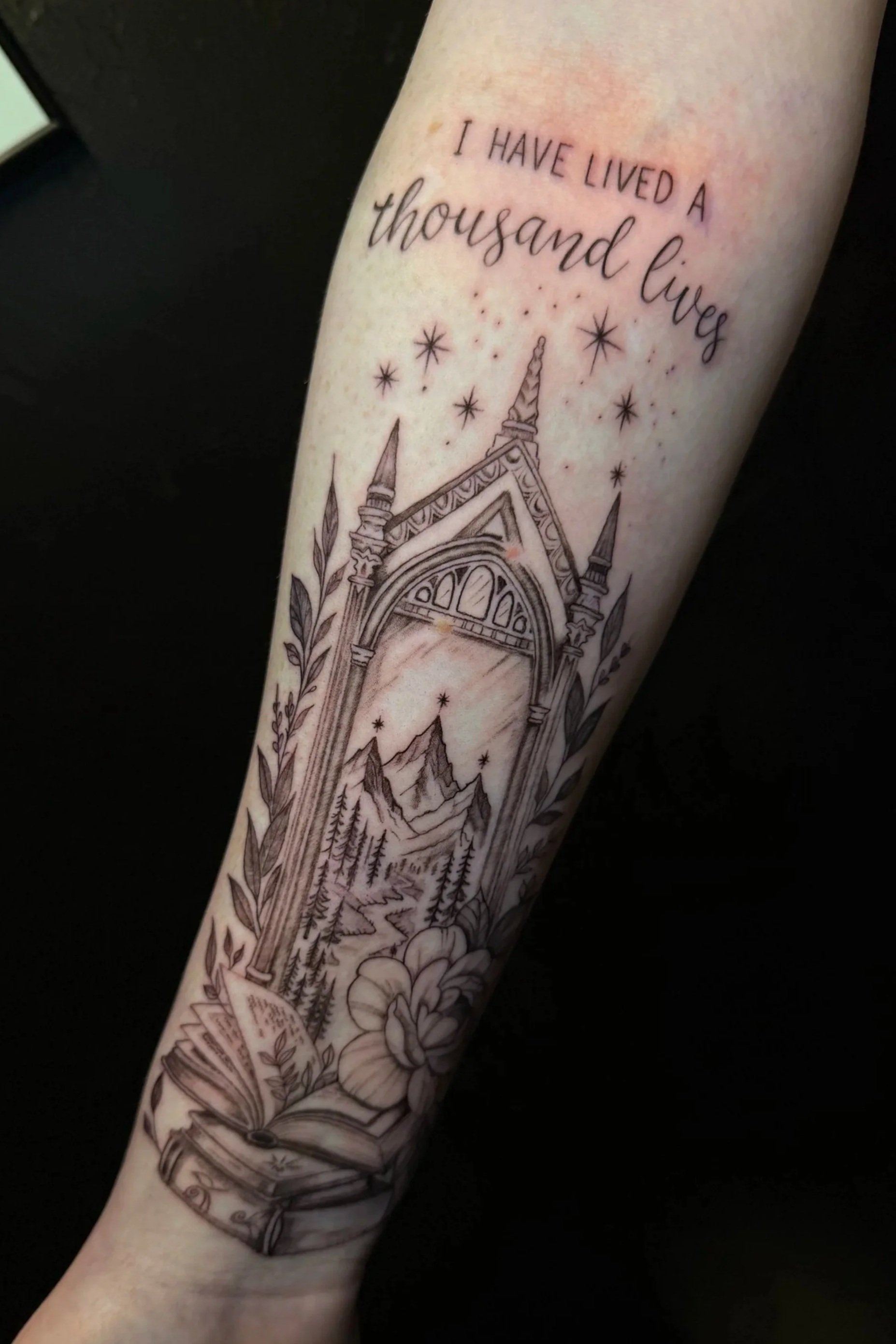 Tattoo of a mountain landscape with trees, clouds, and stars, featuring an arched gateway with intricate designs, and a quote above that reads 'I HAVE LIVED A thousand lives'.