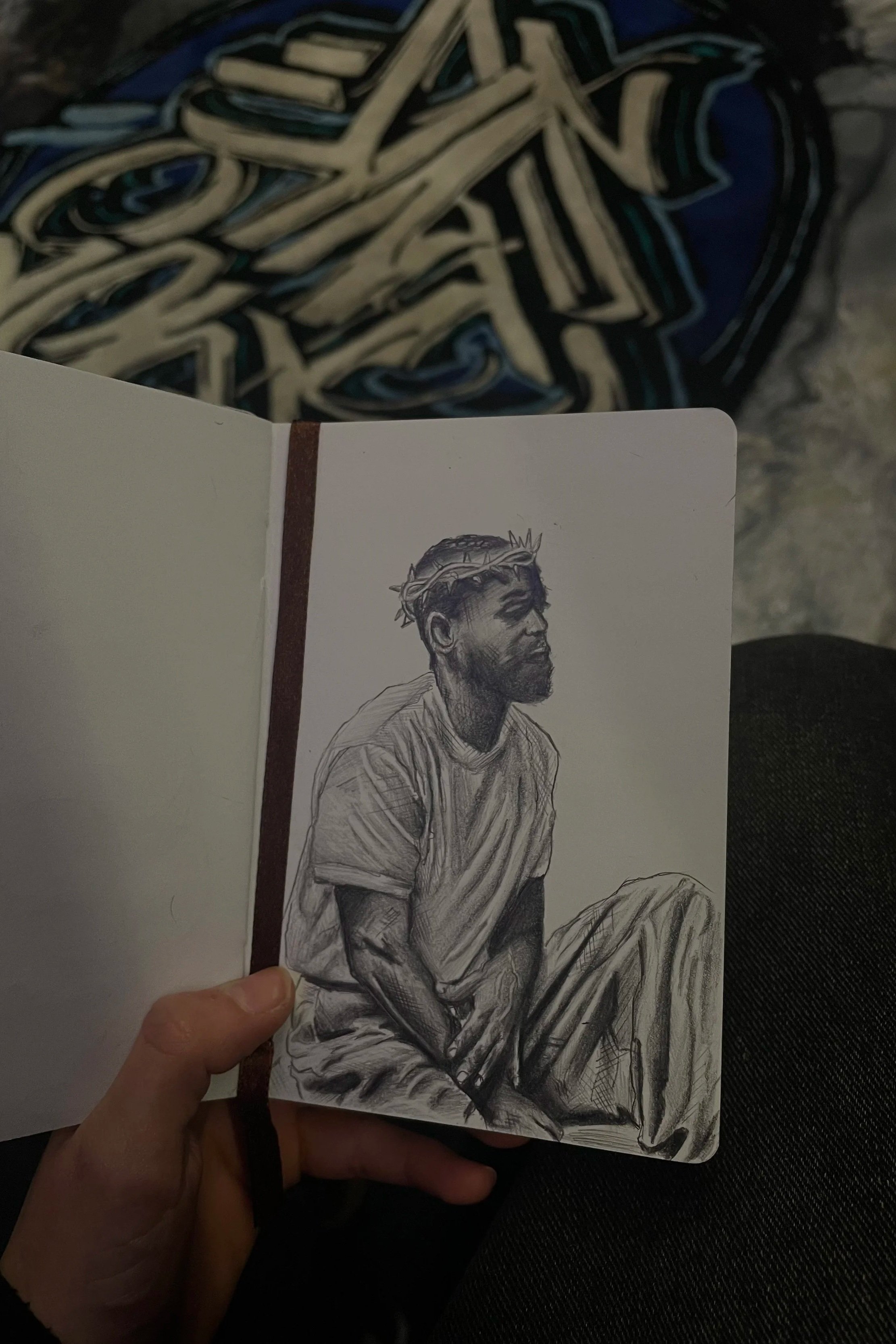 A detailed black and white sketch of a man with a beard and a crown of thorns on his head, wearing a simple shirt, sitting with his arms resting on his legs.