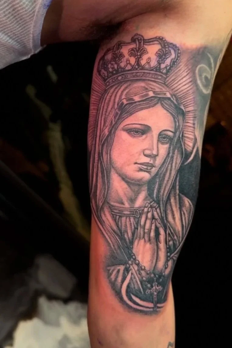 Tattoo of Our Lady of Guadalupe on a person's arm, depicting the religious icon with a crown, praying hands, and a rosary.