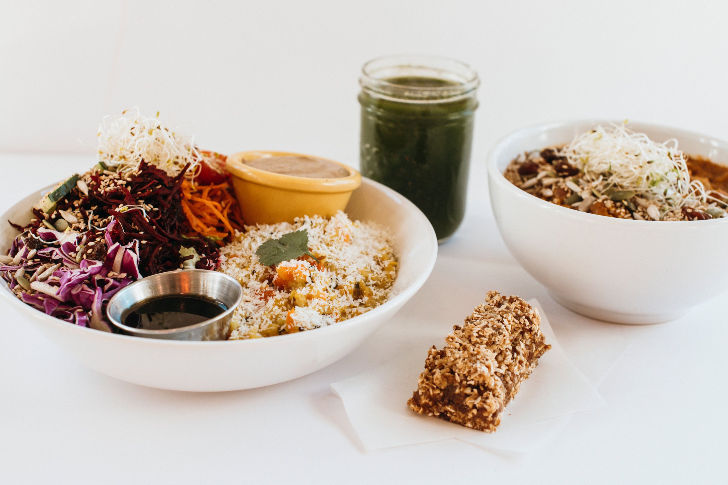 Assorted vegetarian dishes with salads, a mountain of shredded vegetables, a bowl of chili with topping, a drink, and a granola bar on a white surface.