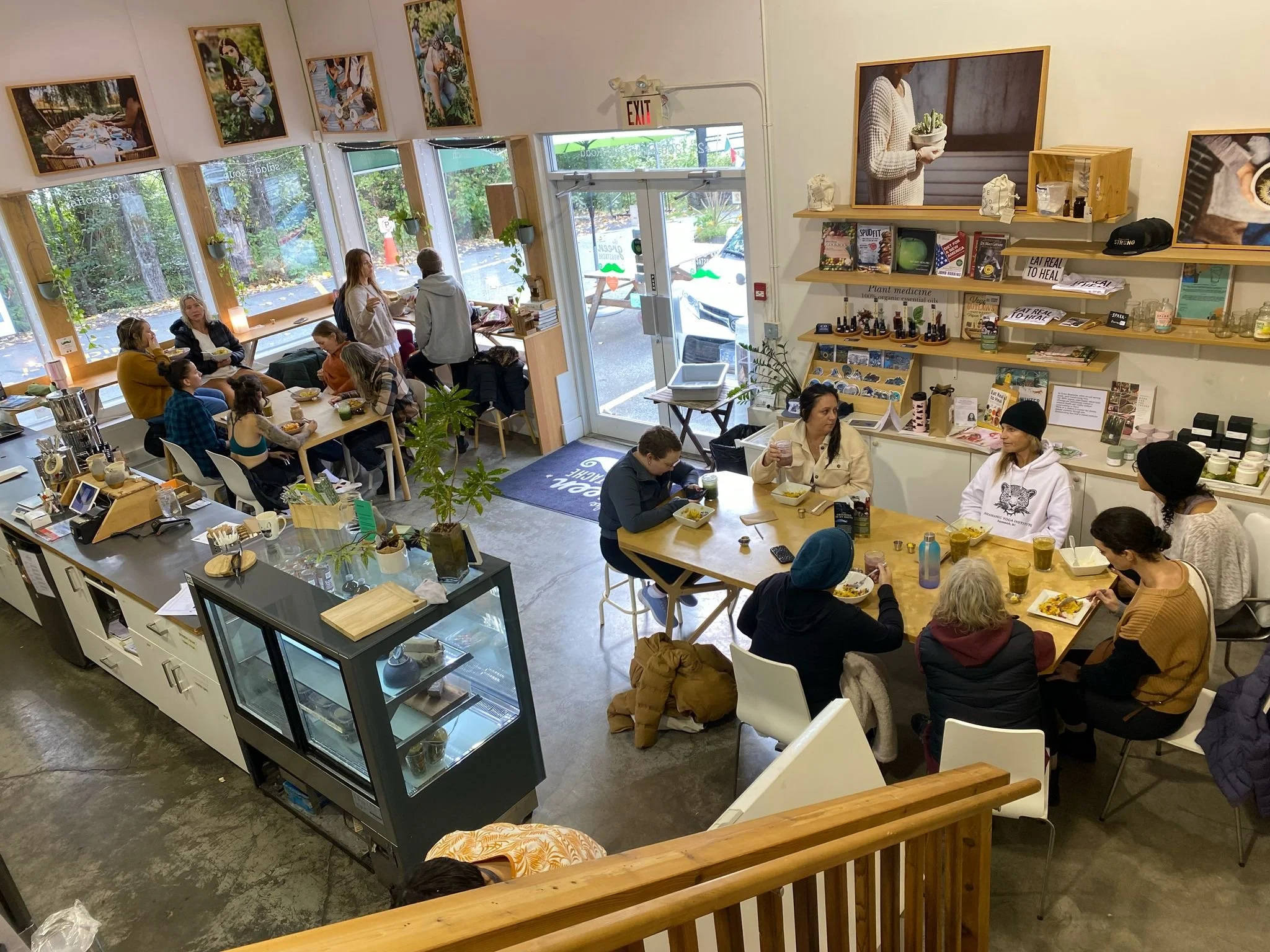 A busy coffee shop with customers dining and socializing, large windows with outside greenery, artwork on the walls, and a display case of baked goods.