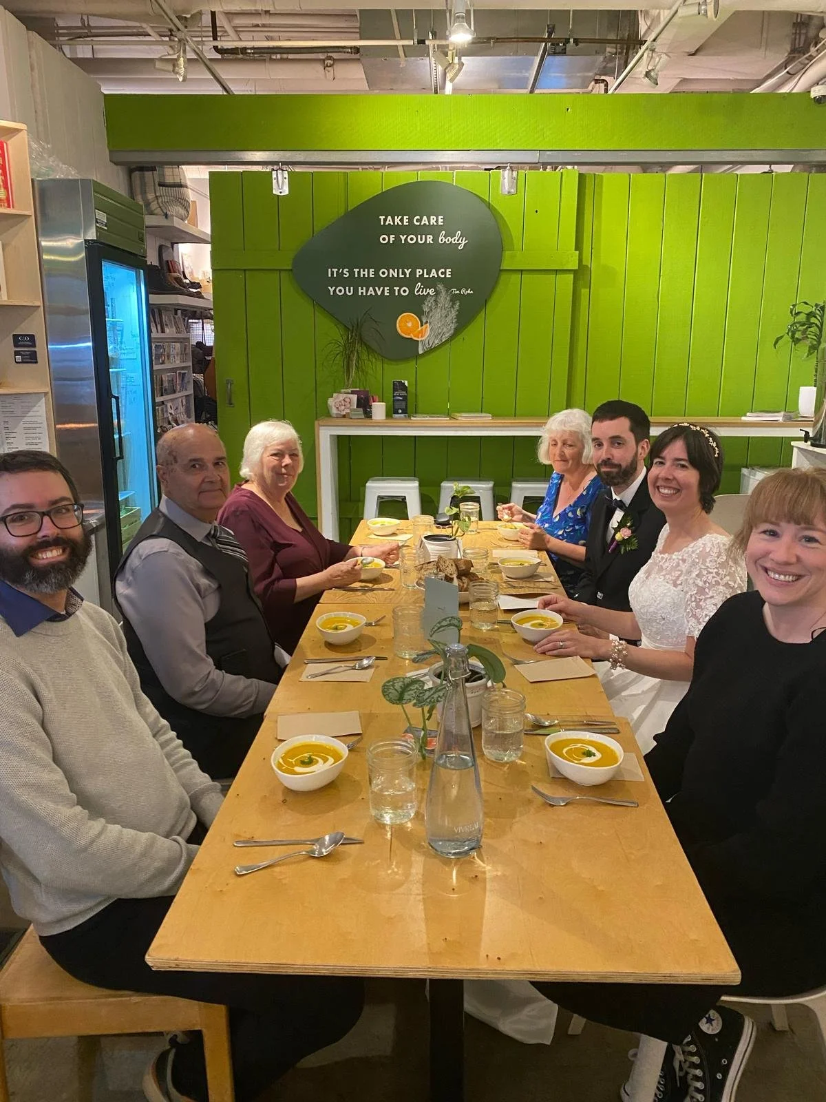 A group of people sitting around a dining table, enjoying a meal in a restaurant with green interior decor and a sign on the wall that reads, 'Take care of your body. It's the only place you have to live.'