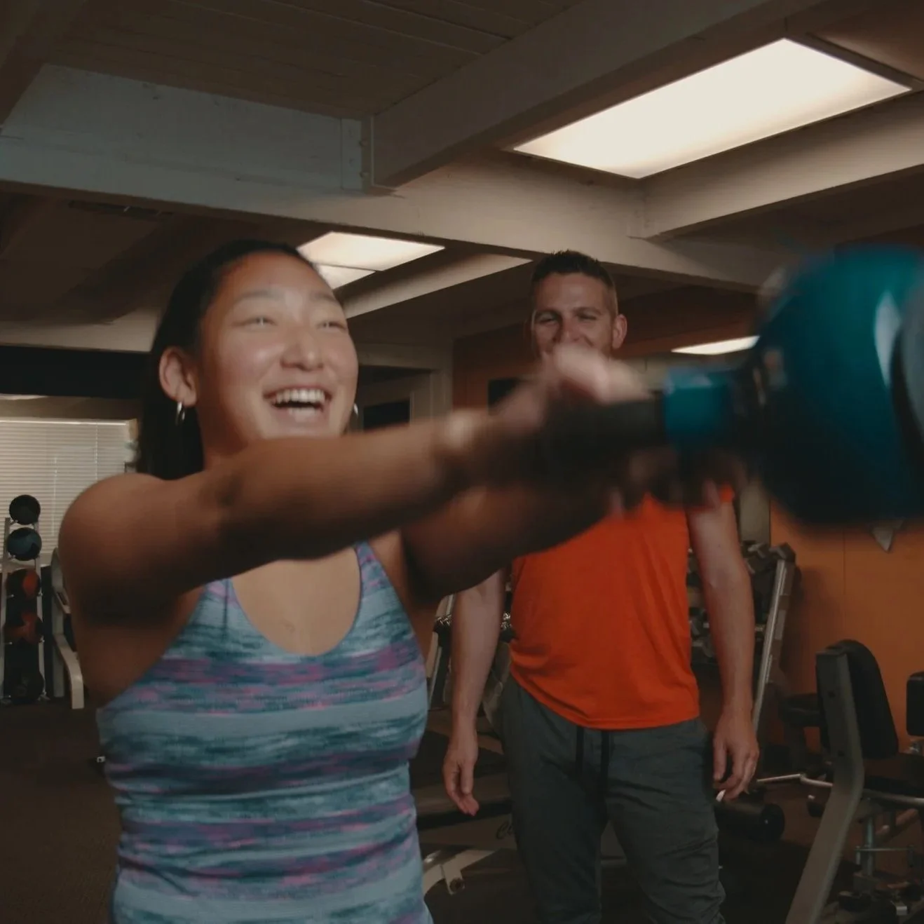 A woman laughing and holding a blue and black drill in a gym or fitness center, with a man in an orange shirt smiling in the background.