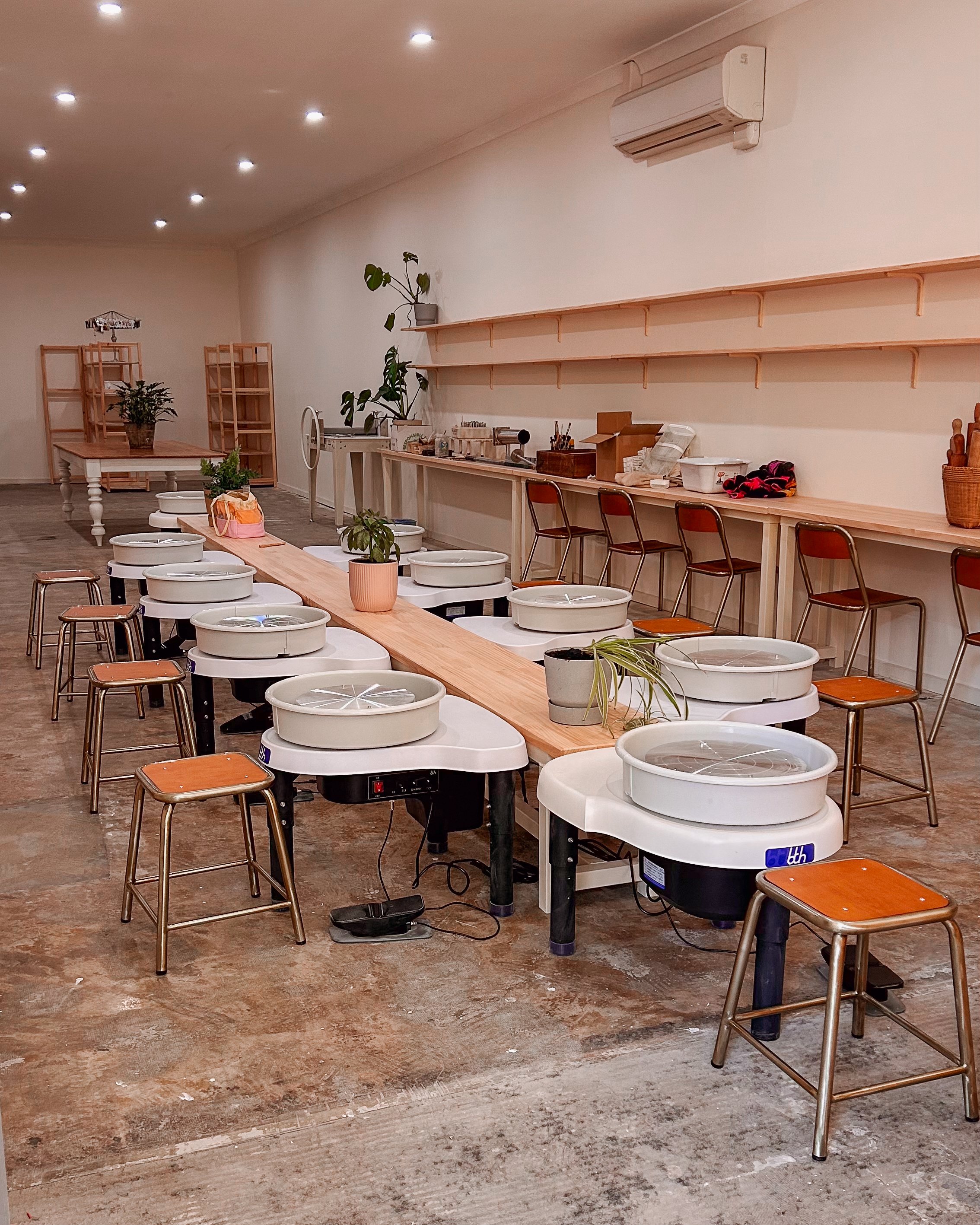 A row of pottery wheels with brown stools in a ceramics studio, with shelves, plants, workstations and a white wall with an air conditioning unit.