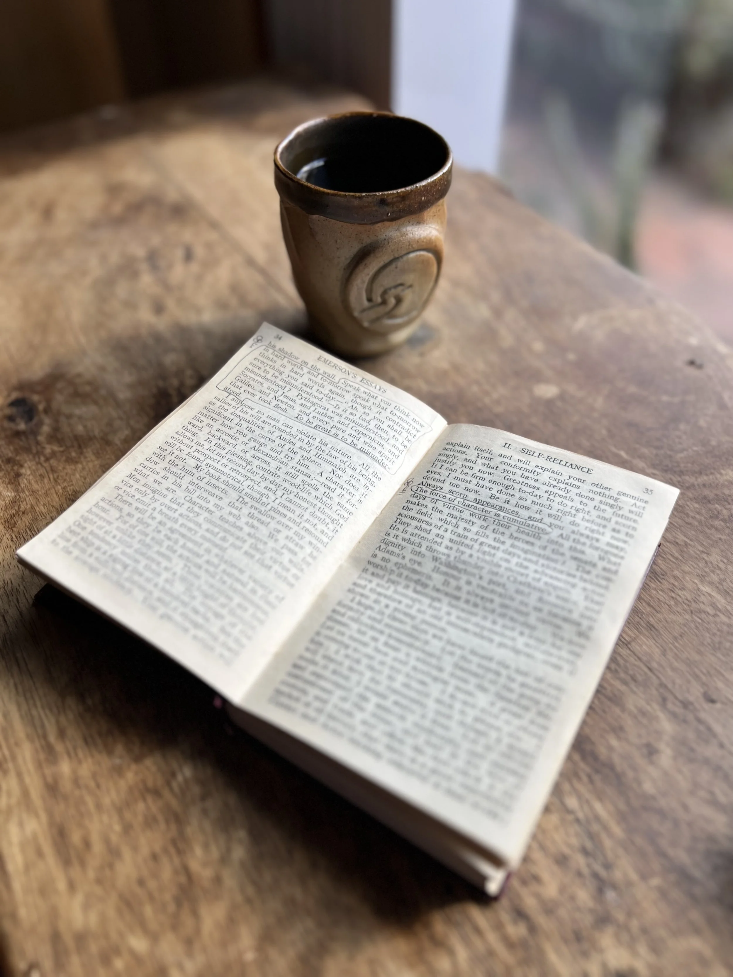 Open page of Emerson Essay on Self-reliance discussing consistency and being misunderstood, tea cup in background.