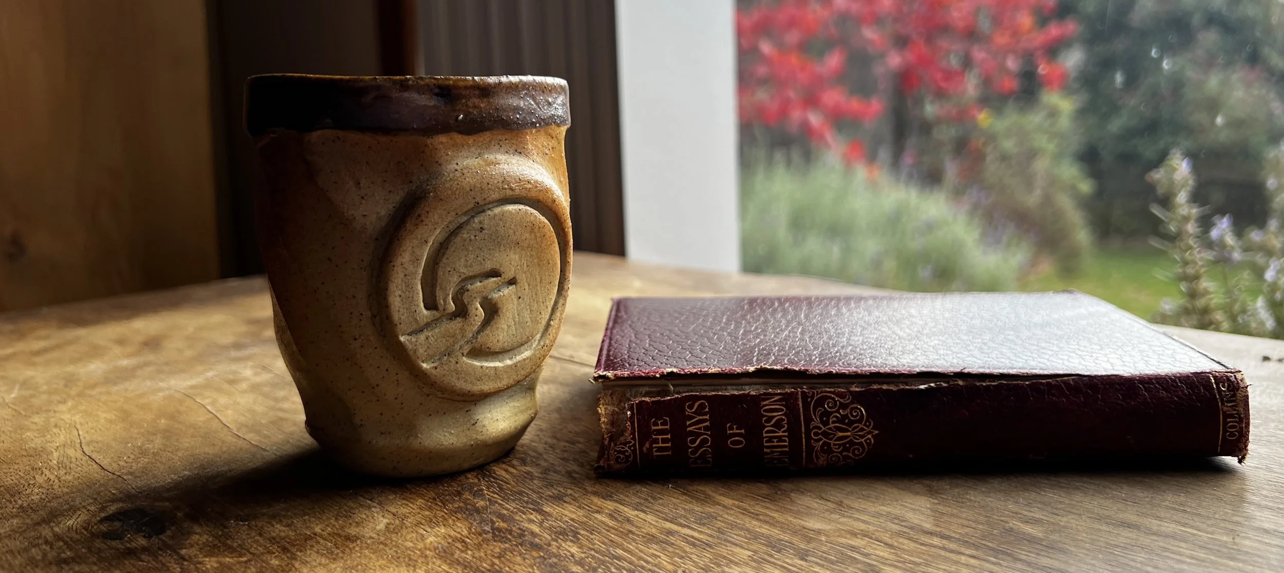 Image of closed book of Emerson's Essays, tea cup and autumn leaves outside of window.