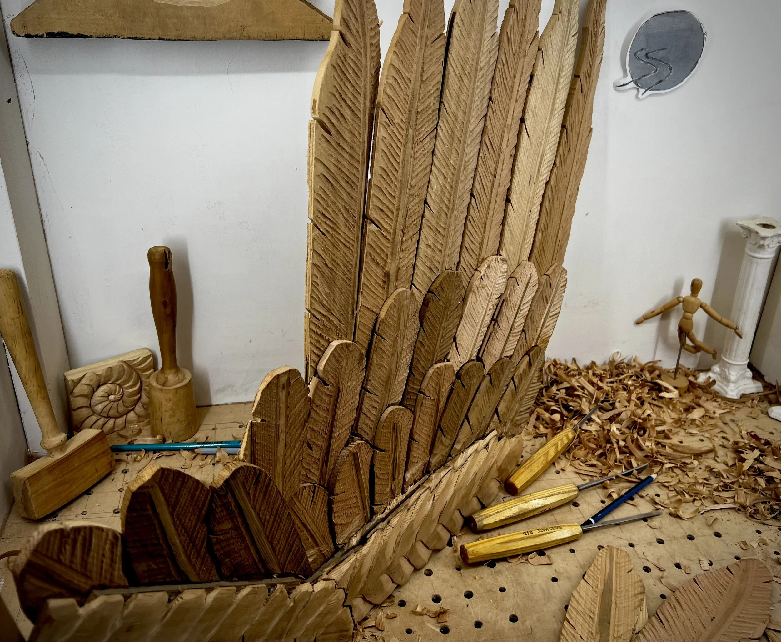 Hand-carved wooden angel wing in progress on a workbench, unfinished feathers, chisels and cedar shavings visible