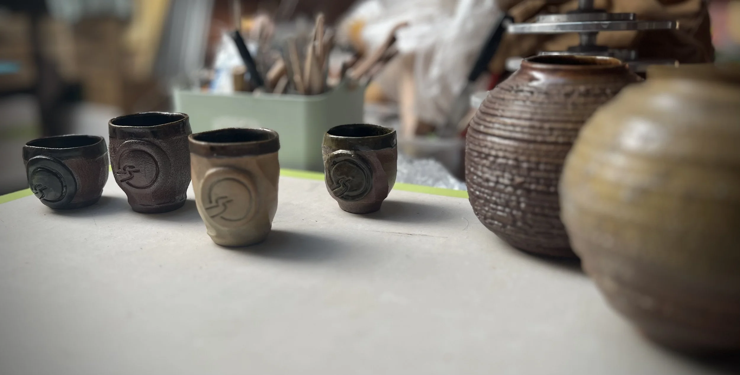 Four hand made clay cups with the Insightful path logo, resting on a table near pots and potters tools.