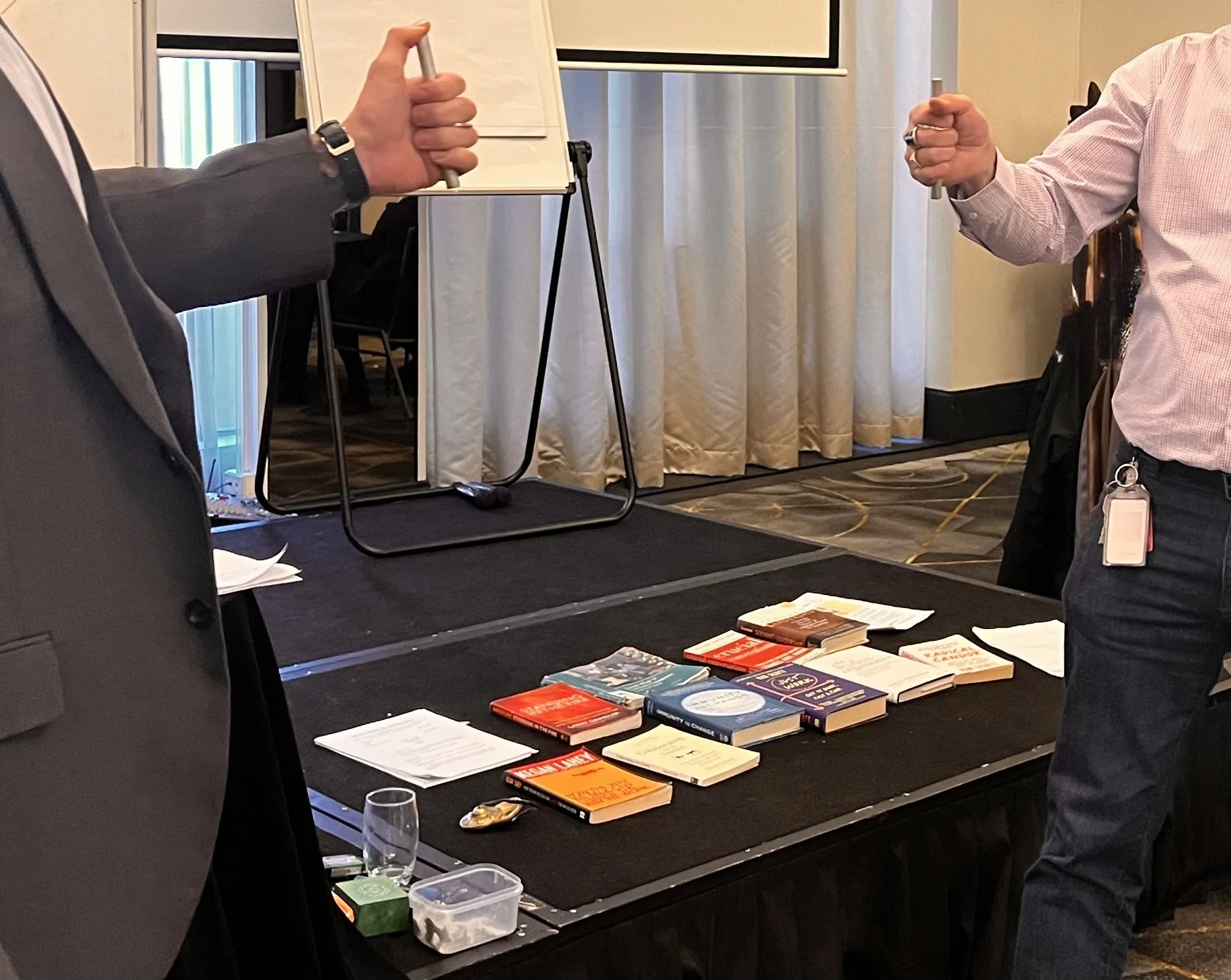 Two people pulling a fine fishing line to test where a knot will break, with books visible in the background, representing standards, limits, and accountability.