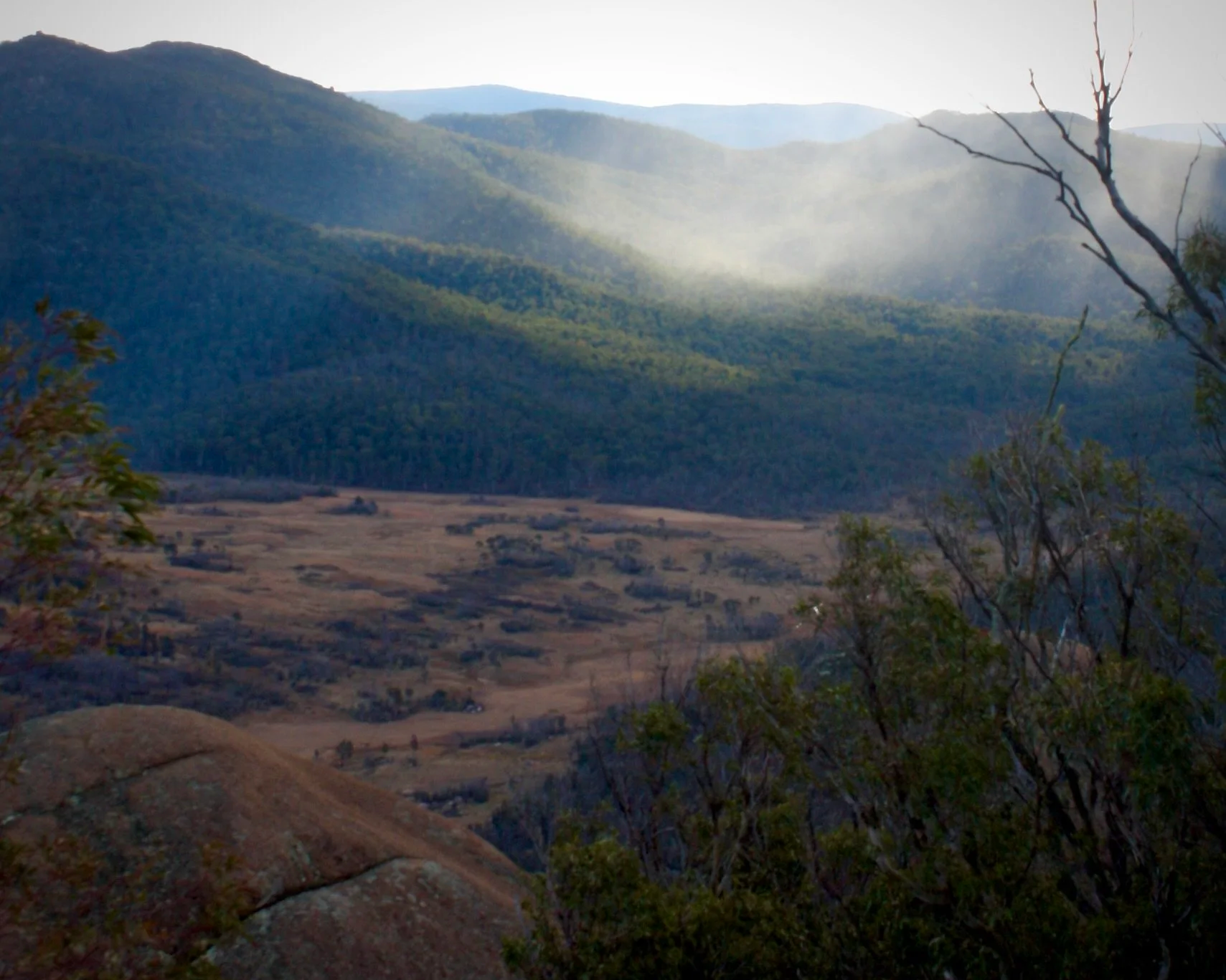 A wide Australian landscape showing rain and sunlight illuminating distant valleys, symbolising perspective, emergence, and shared inquiry across space.
