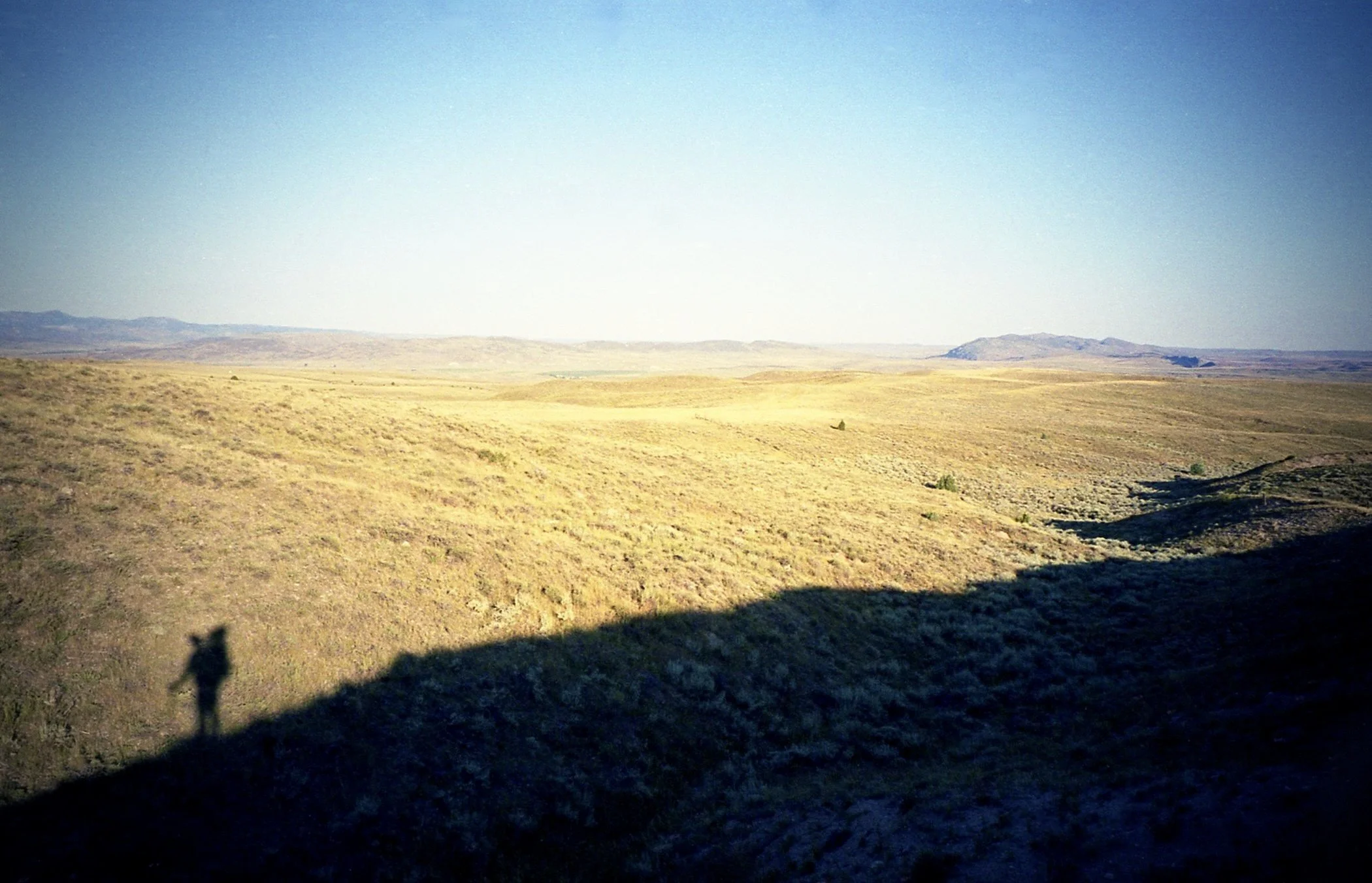 A silhouette of a man with a backpack and walking sticks against a dry high alpine desert hill and mountains in the distance.