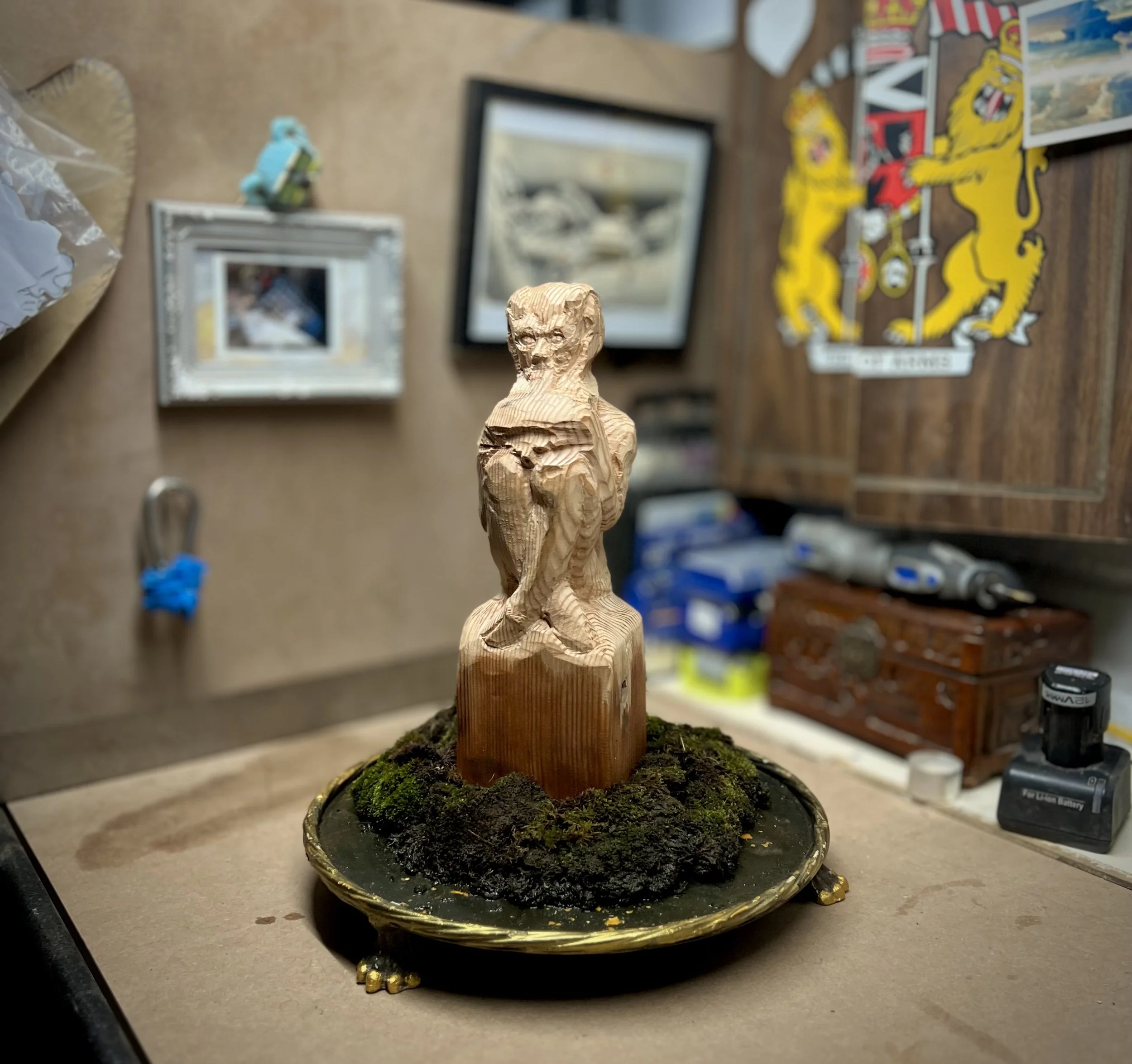 Nearly finished cedar wood Pan sculpture seated cross-legged on a stump, resting on a golden claw-footed-platter, nestled in a bed of living moss.
