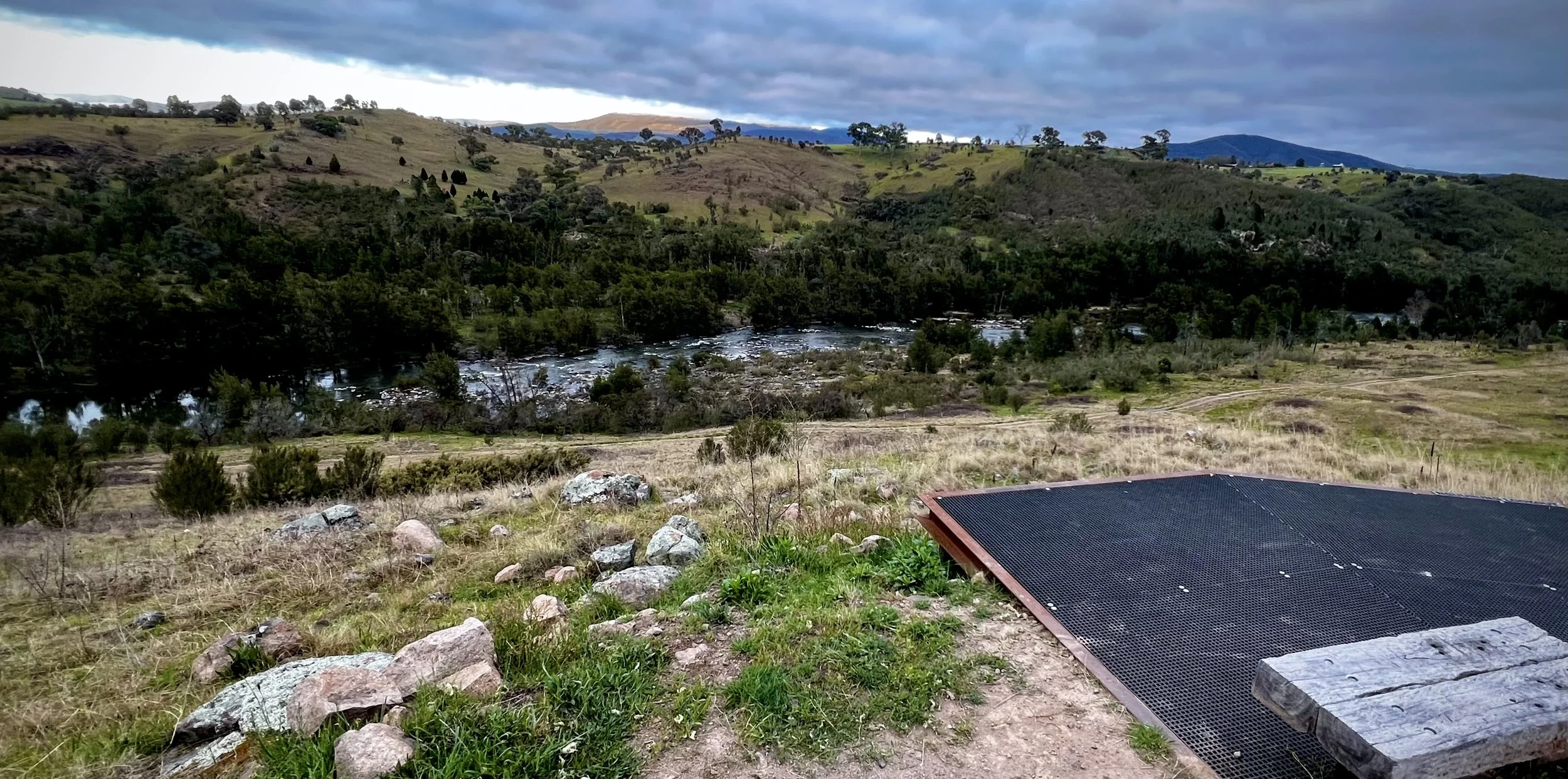 A timber bench on a metal lookout platform above a tree-lined river, with grassy hills beneath a cloudy sky.