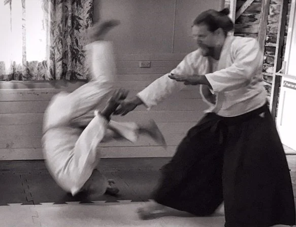 Black Belt, James Samana, practising Aikido, mid-technique, throwing another practitioner while maintaining focus and balance, illustrating dynamic tension and embodied learning.