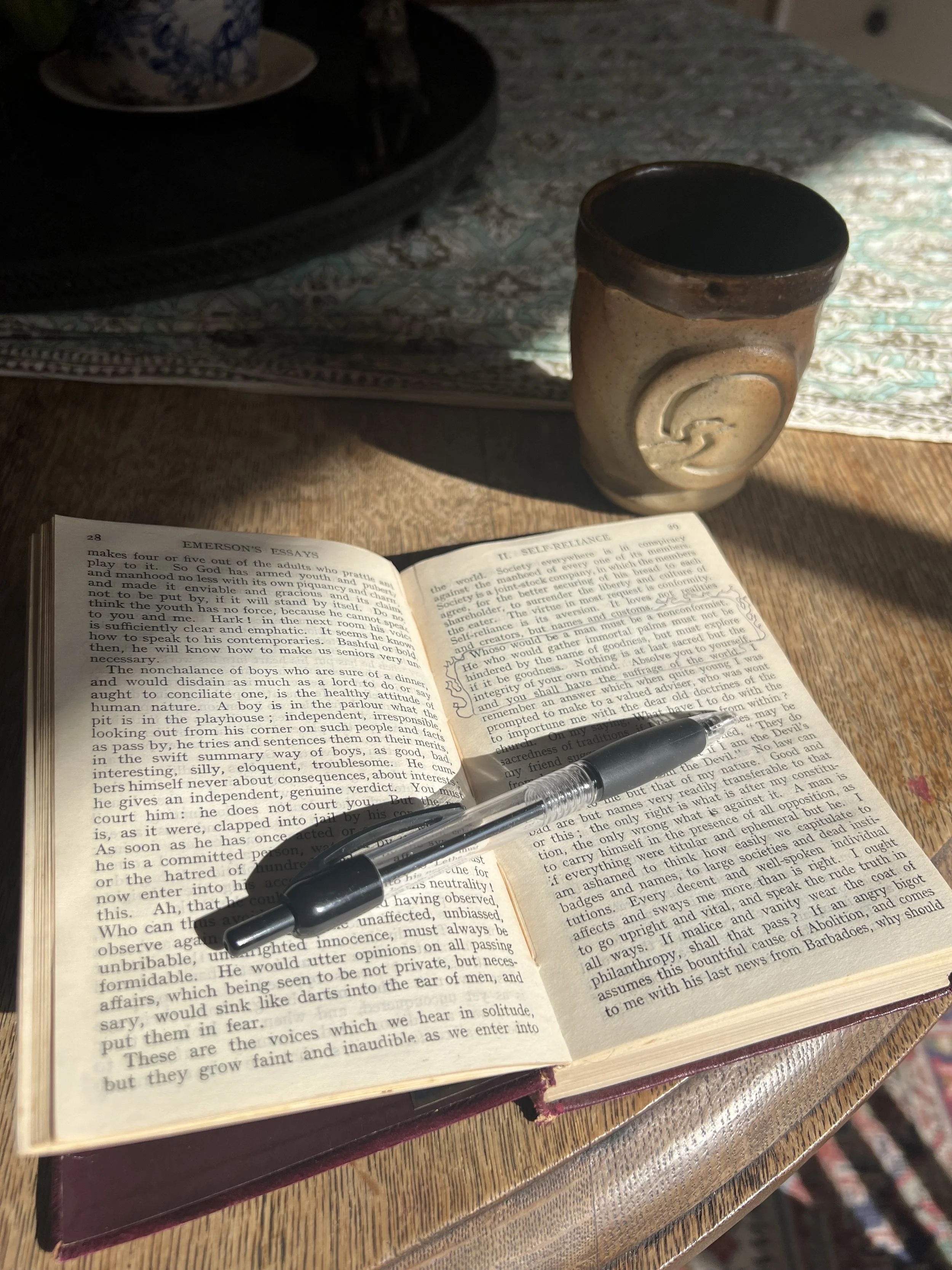 Emerson Essay open to nonconformity quote on wooden table in sunlight, teacup in background
