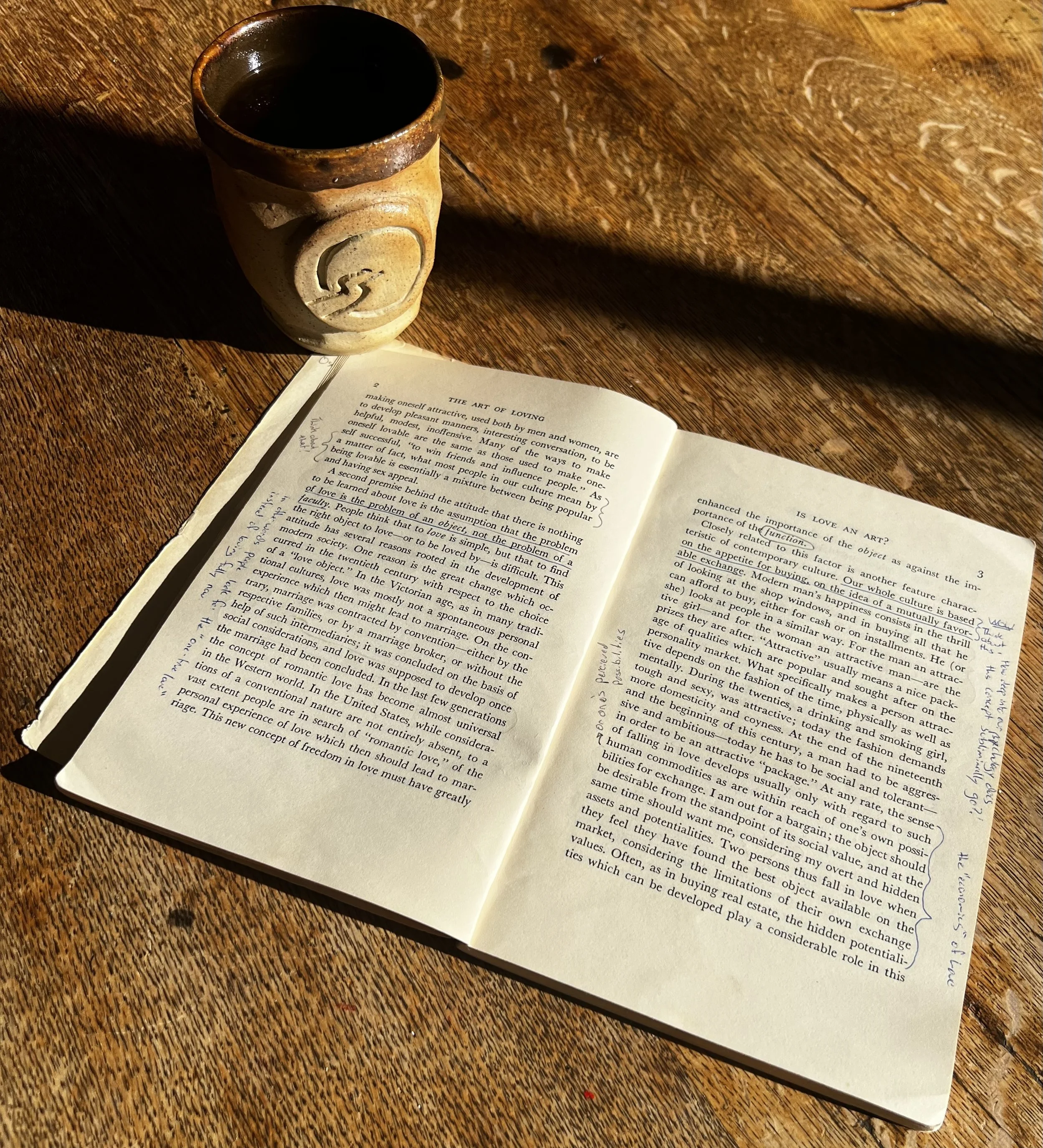 Open copy of The Art of Loving filled with handwritten notes beside a handmade clay cup with the Insightful Path logo in morning sunlight.