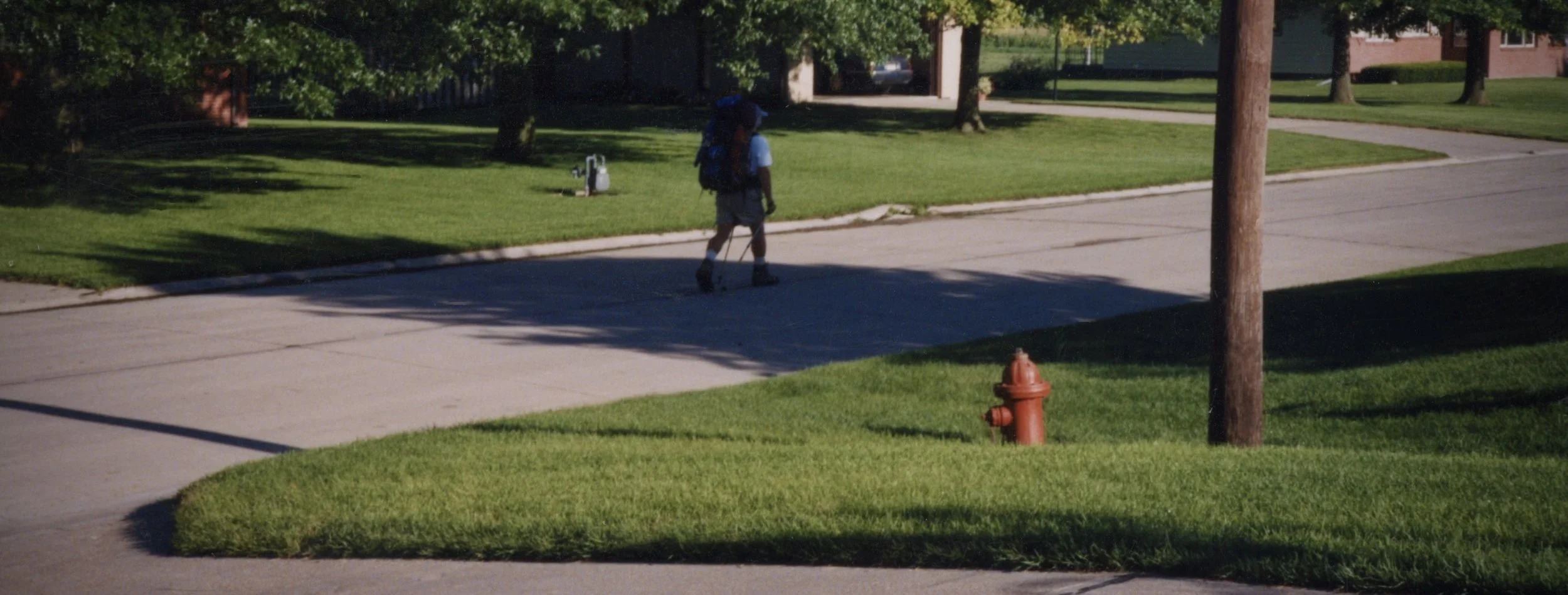 Man walking down a quiet suburban street lined with red brick houses and green lawns during late afternoon light.
