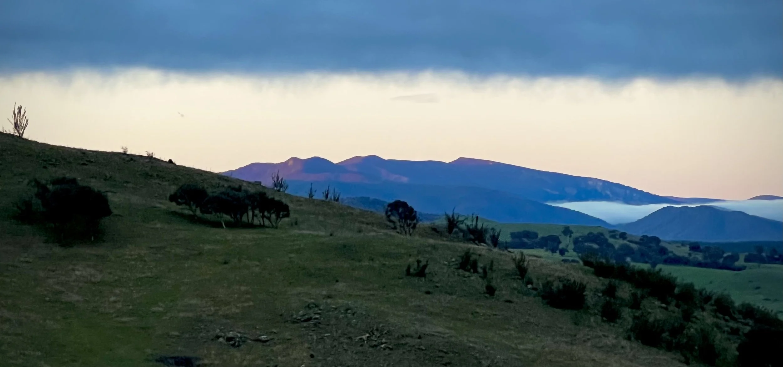 Rolling hills north of Canberra beneath dark clouds, with a pale band of light above the horizon and mist between distant ridges.