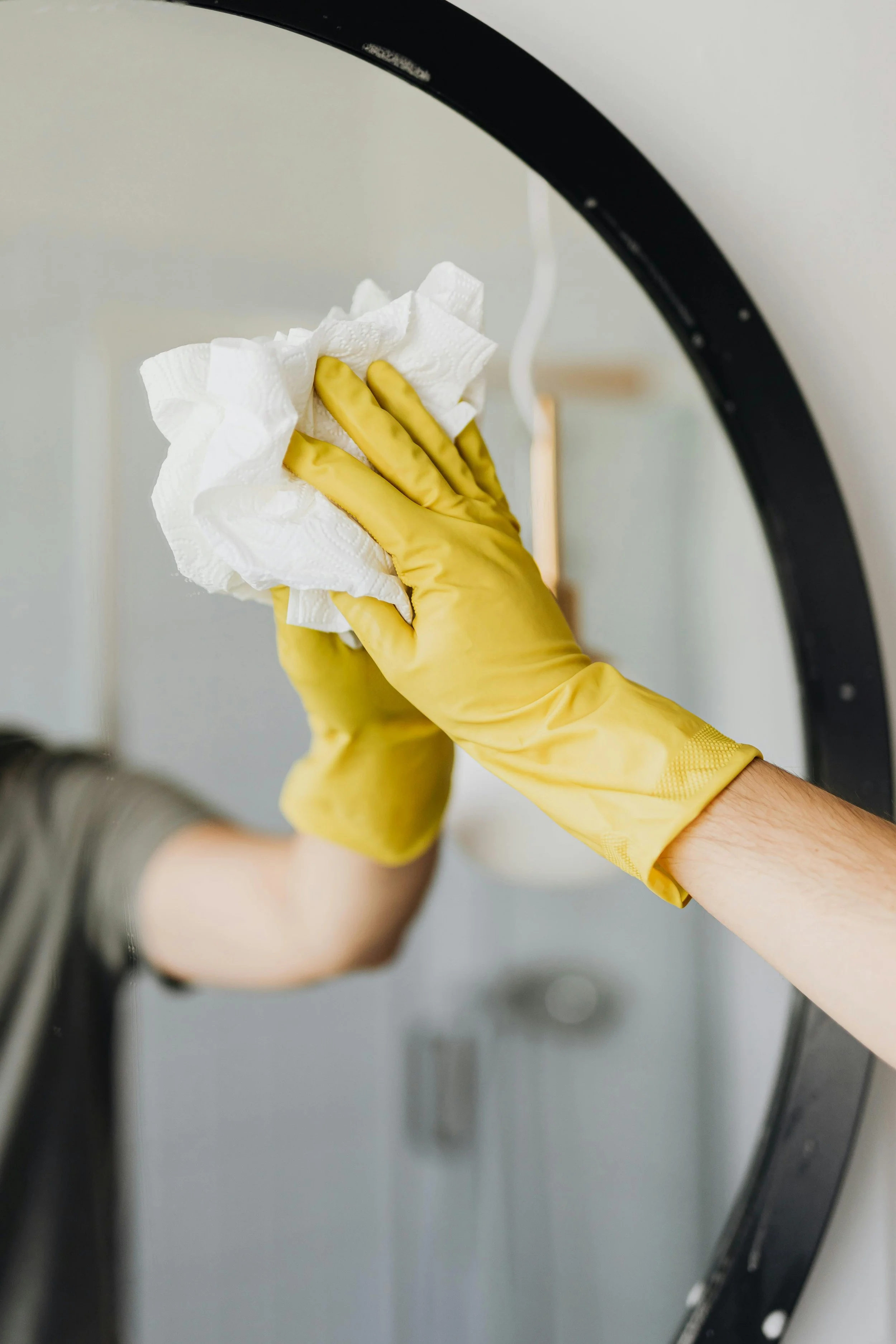 Person cleaning a round mirror with a white cloth using yellow rubber gloves.