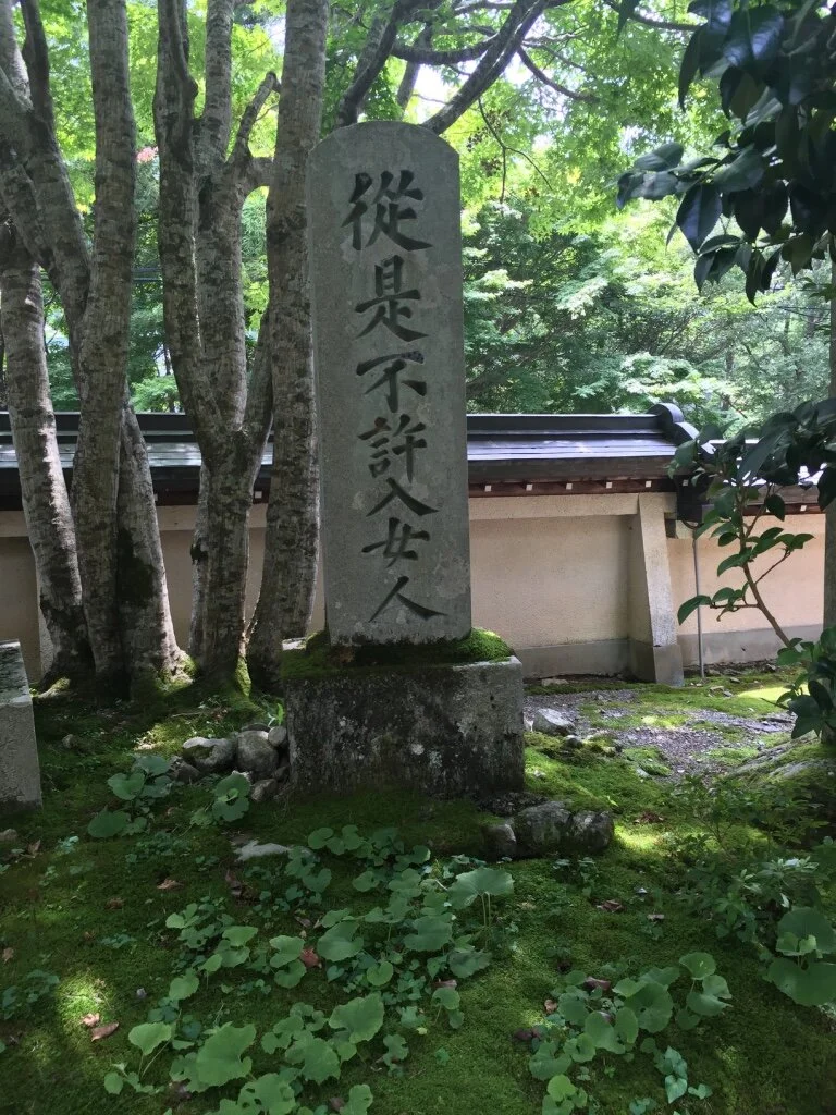 A stone monument with Japanese kanji characters inscribed, surrounded by trees, moss, and greenery in a tranquil outdoor setting.