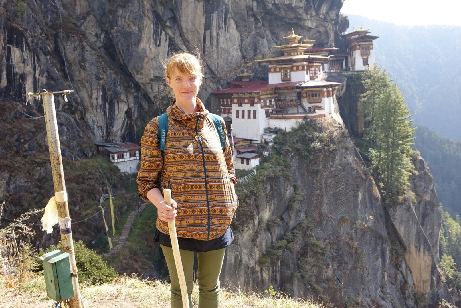 A woman with a backpack and walking stick standing on a trail with Tiger's Nest Monastery in Bhutan in the background.