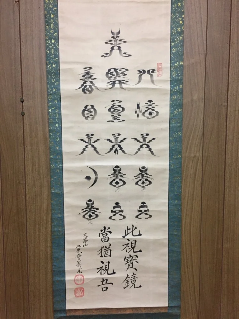 Traditional Japanese hanging scroll with black calligraphy and symbols, bordered by patterned fabric, mounted on a wooden wall.
