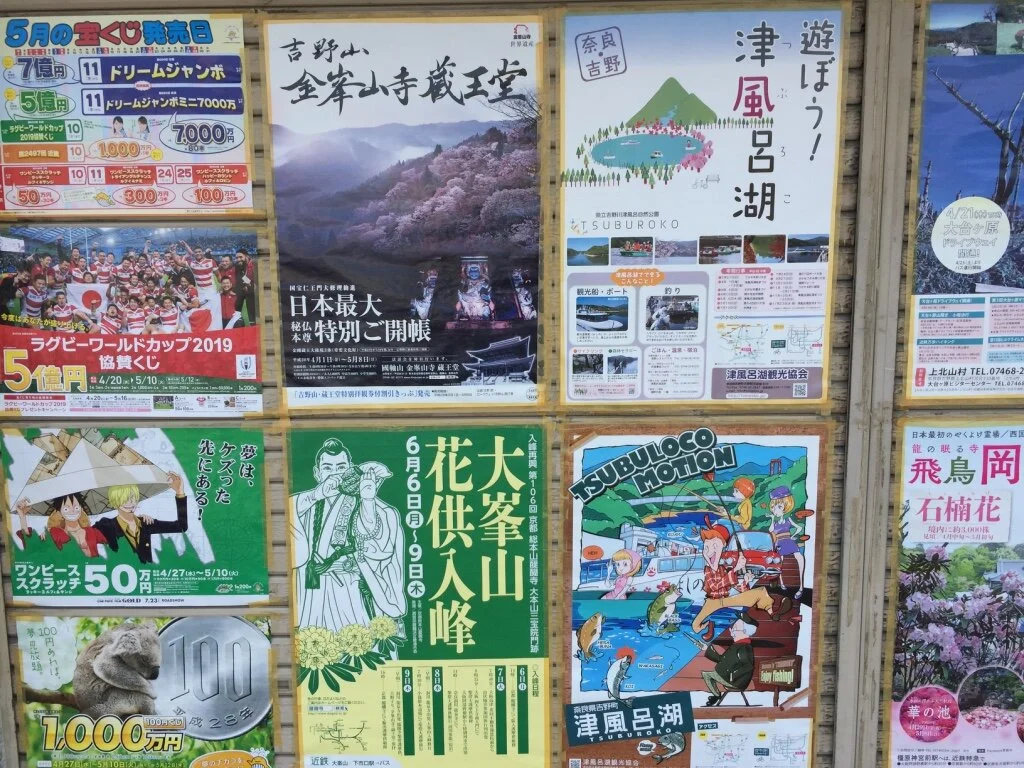 A bulletin board with various colorful posters, including advertisements for local events, a rugby cup, a special event at a temple, promoting the Tsuburoko Lake, and scenic attractions featuring mountains, cherry blossoms, and local culture.
