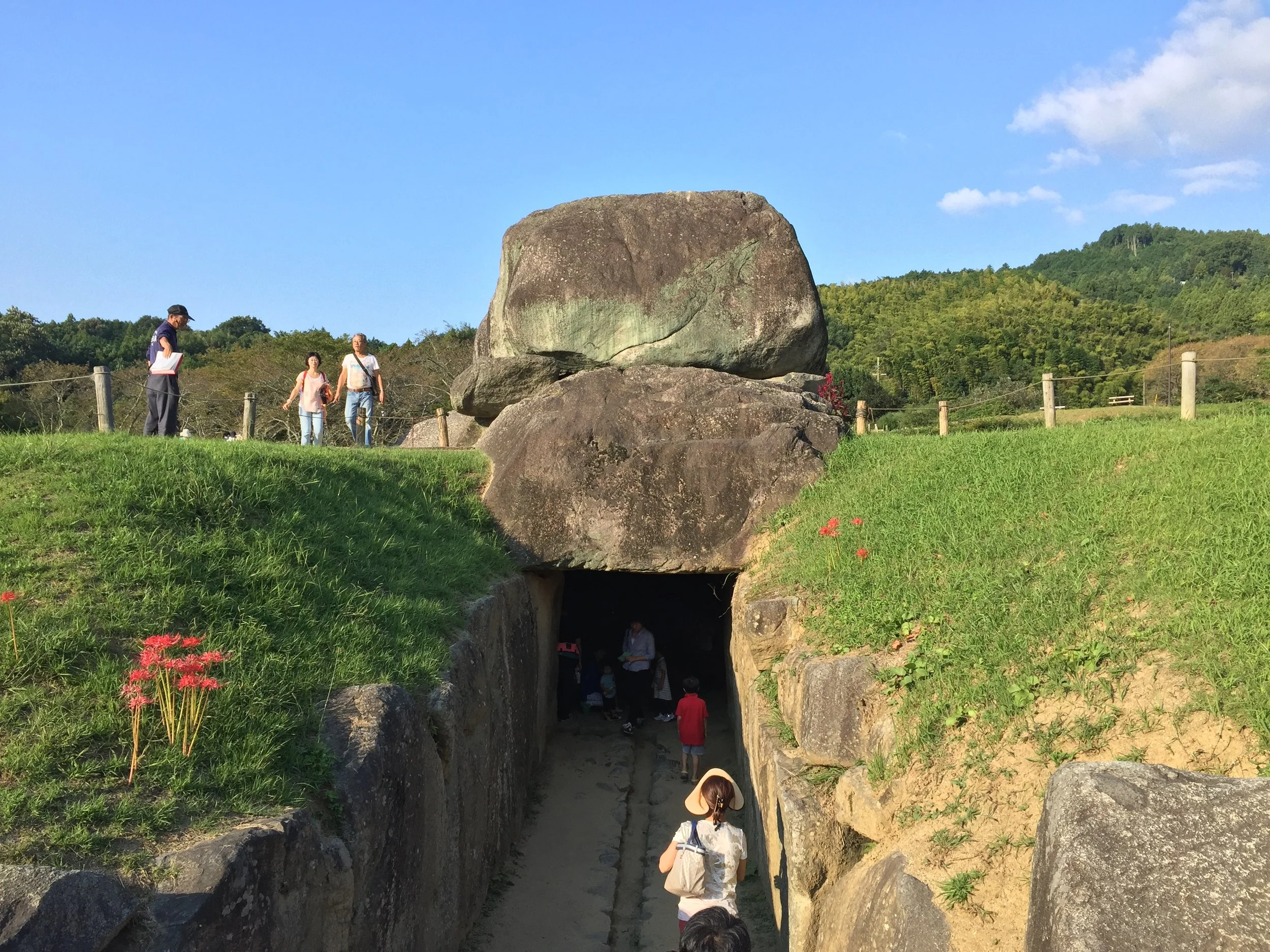 People walking through a tunnel beneath large rocks in a grassy area with hills and trees in the background.