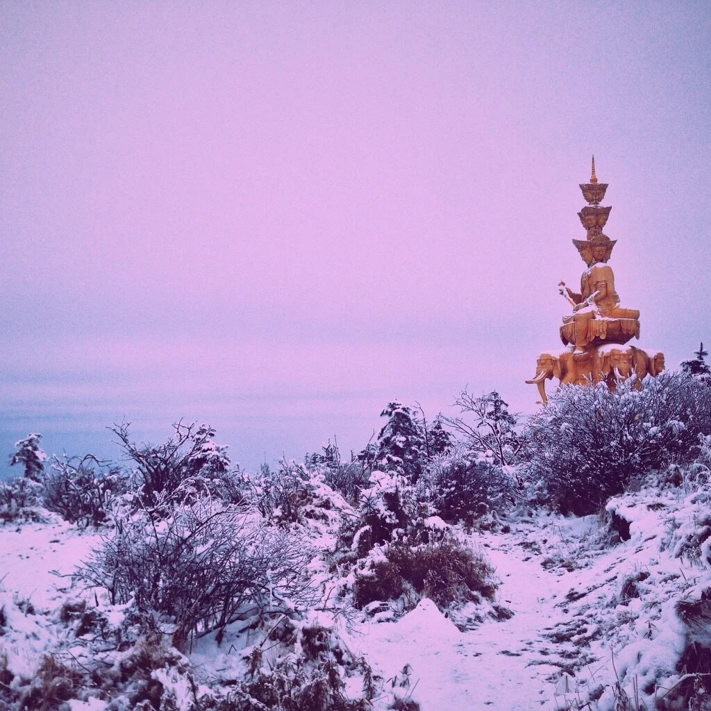Snow-covered bushes and trees with a tall, ornate, multi-tiered golden pagoda-style monument in the distance against a clear purple sky.