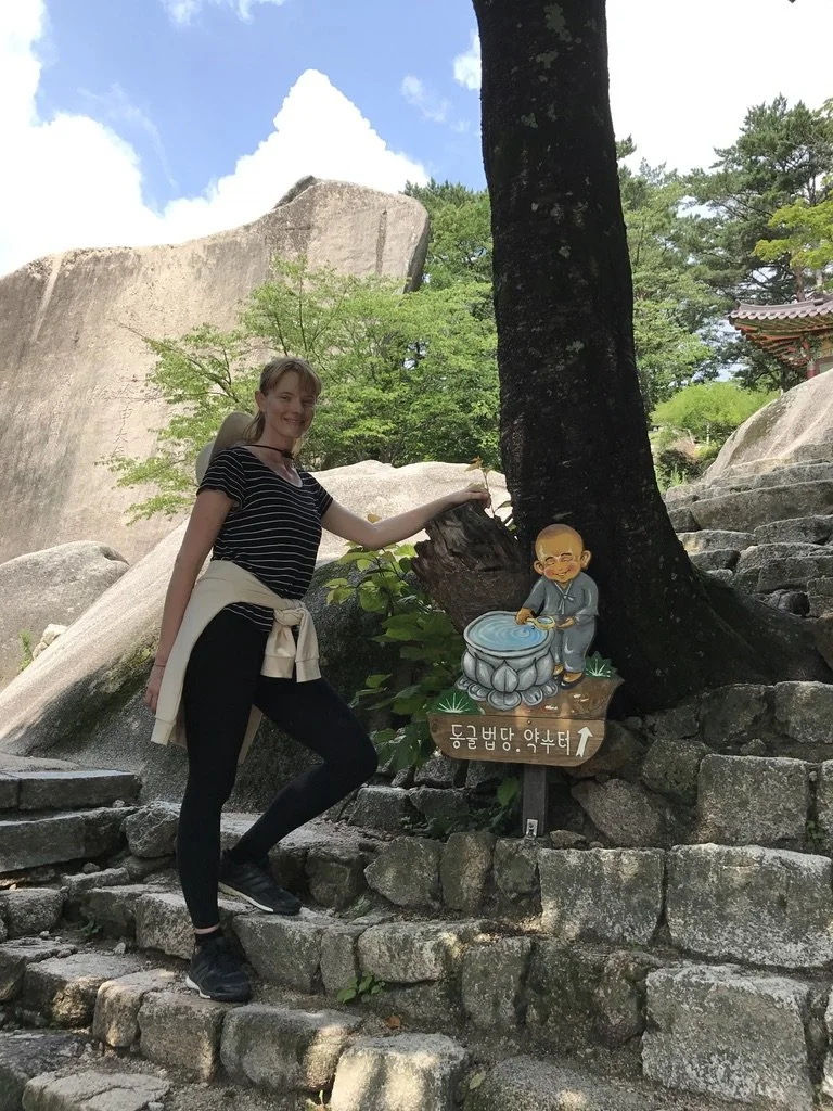 A woman with a beige backpack standing outdoors on stone steps, touching a cartoon sign of a monk with a water basin, near a tree, rocks, and traditional Korean temple architecture in the background.