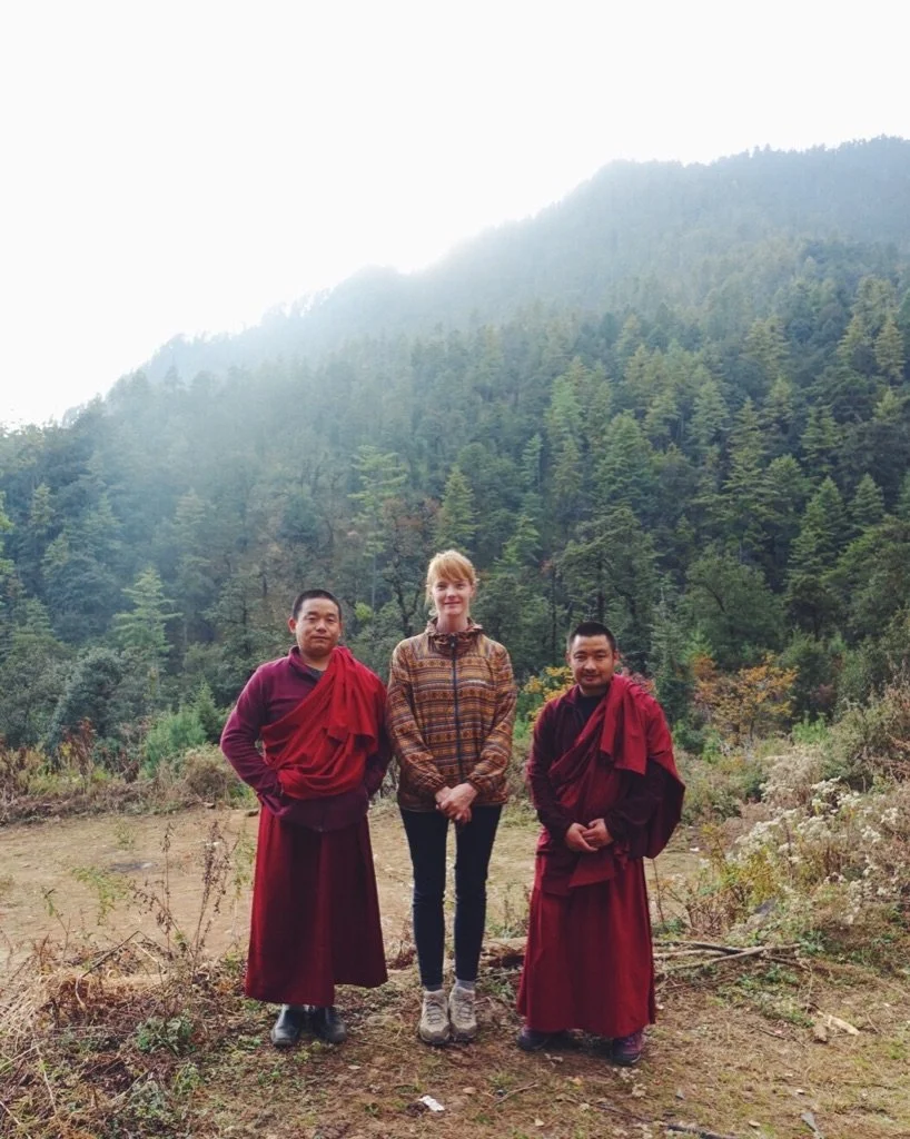 A woman standing between two Buddhist monks outdoors with a forested mountain in the background.