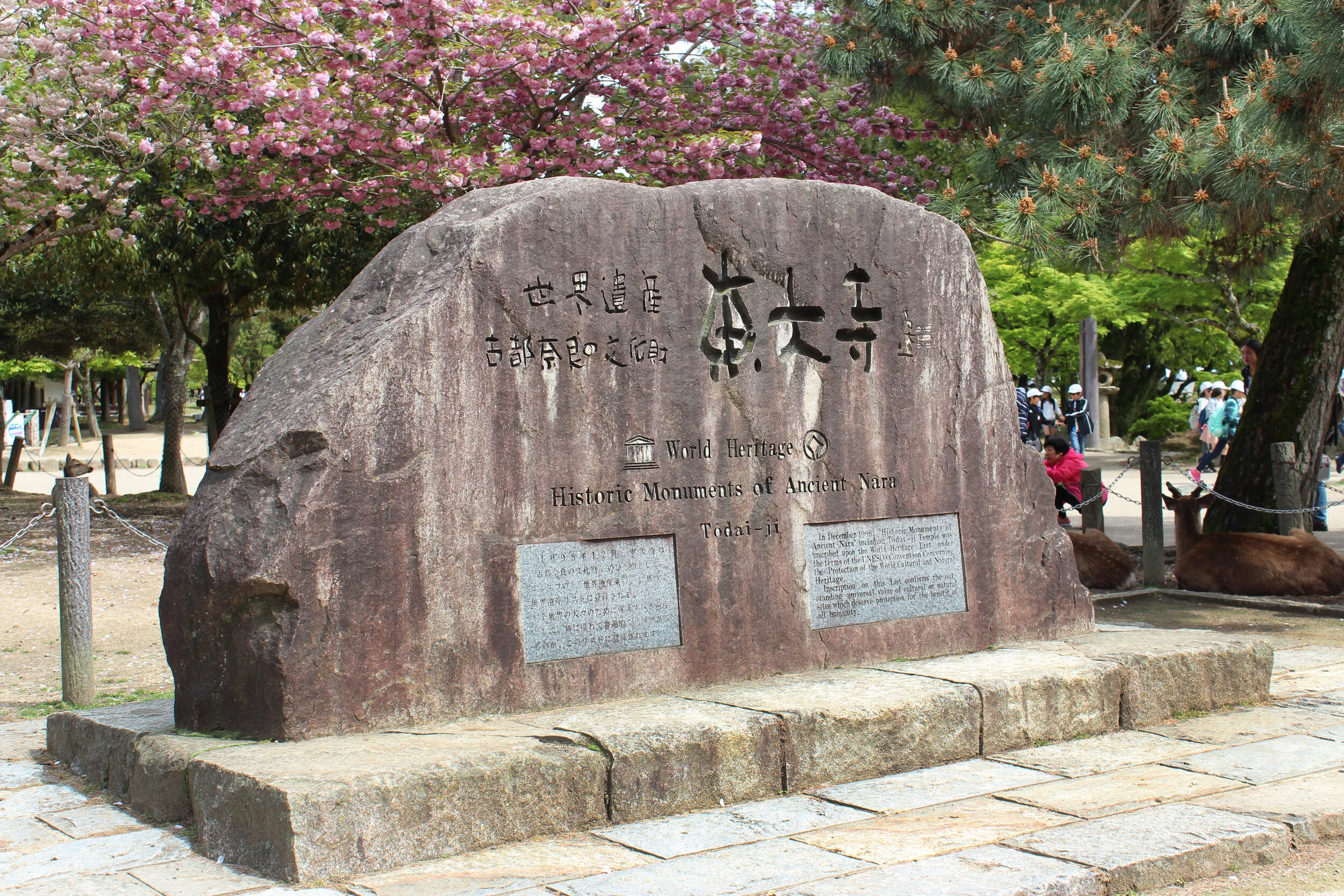 A large stone monument with engraved text, situated outdoors amidst green trees and pink blossoms, with visitors and deer in the background.