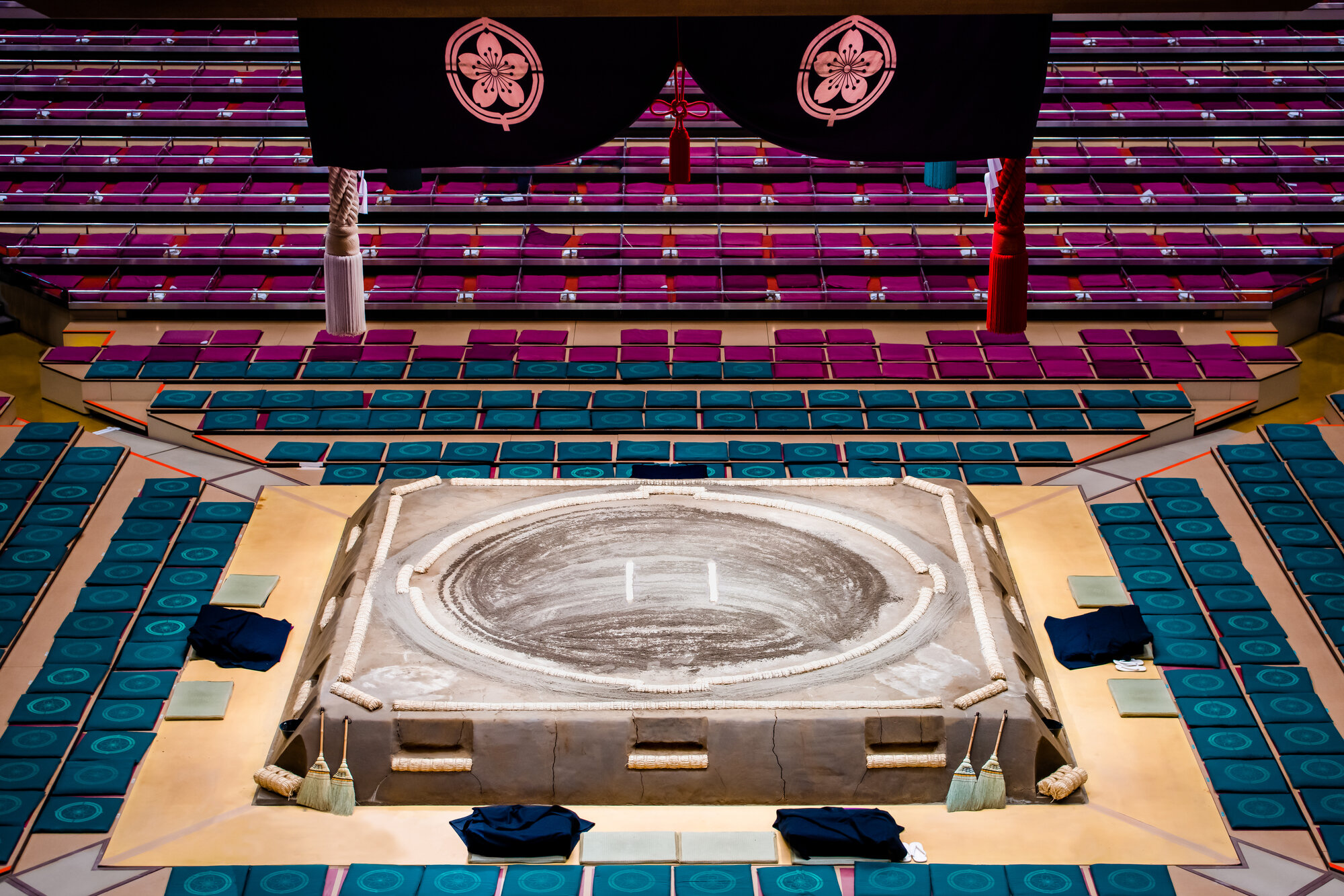 Empty seating area of a traditional Japanese sumo wrestling arena with a raised clay ring in the center, surrounded by cleaning tools and several folded clothes.