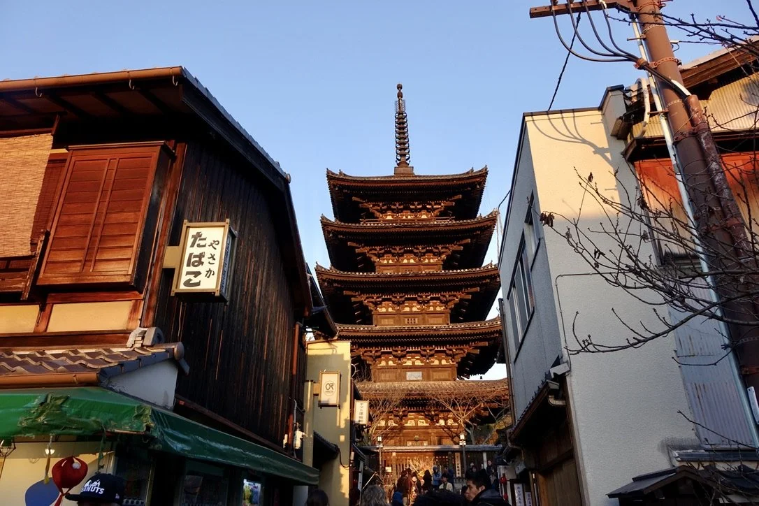 Traditional five-story pagoda with intricate wooden architecture seen between modern buildings on a city street in Japan.